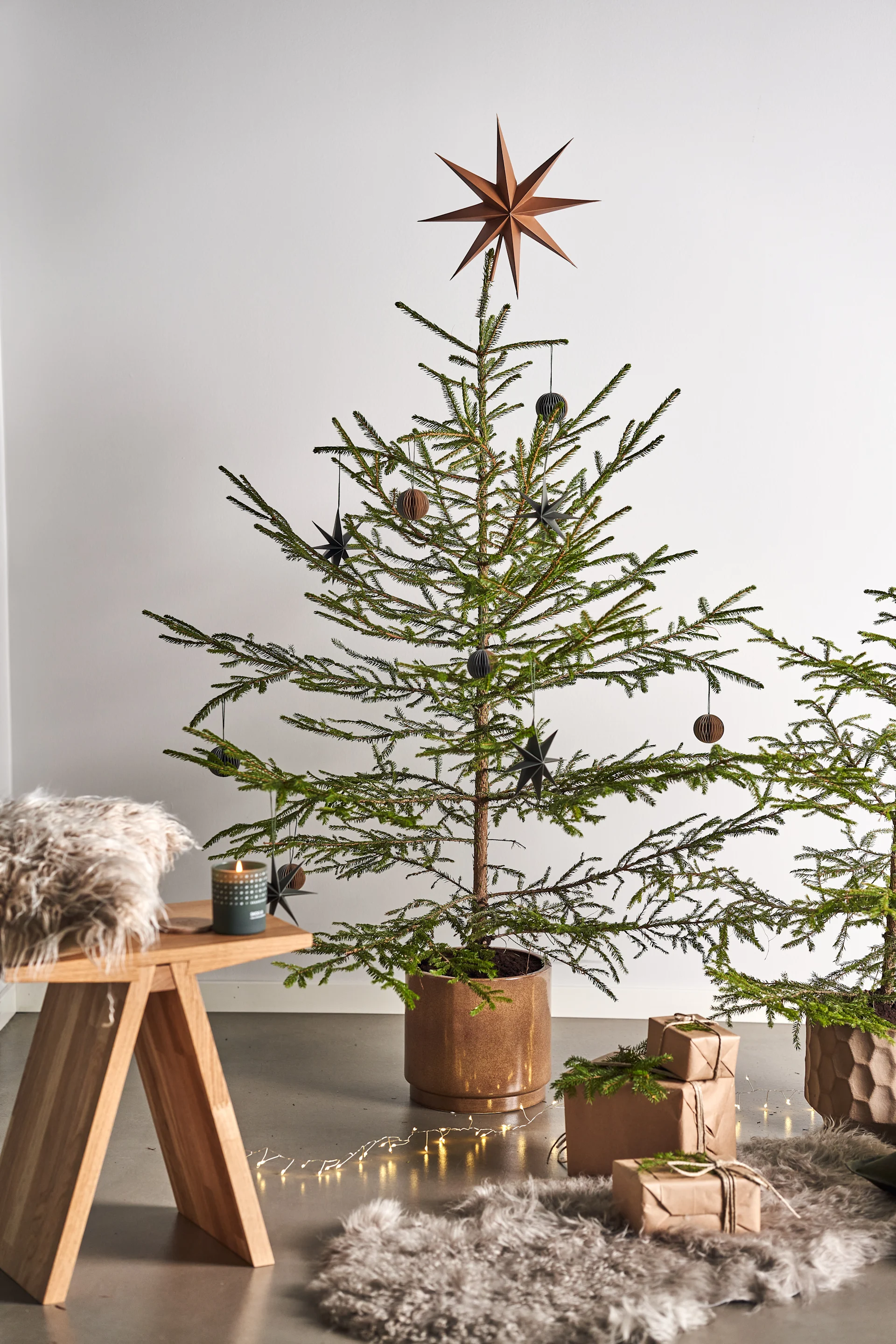Decorate the Christmas tree with Christmas tree decorations for 2021 in 4 different styles according to Nest Trends - Nurture, Share, Boost and Cultivate. Here you see Angle stool from Muubs with A Skandinavisk scented candle in the Forest scent next to packages on a sheepskin rug.