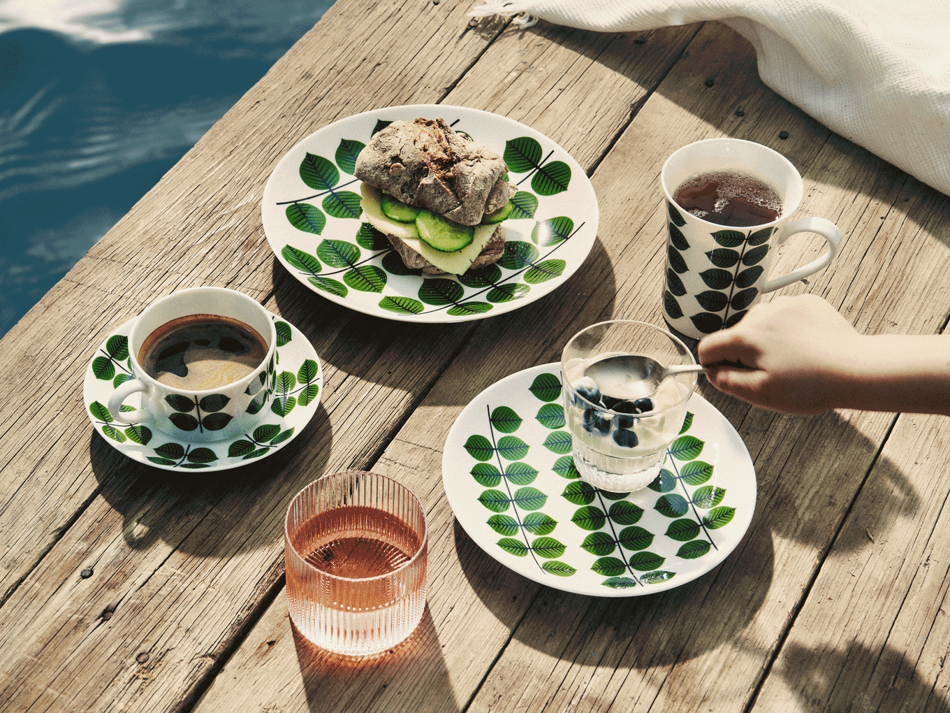 Breakfast on a wooden pier: leaf-patterned plates hold a sandwich, coffee, tea, yogurt with blueberries, and a pink drink.
