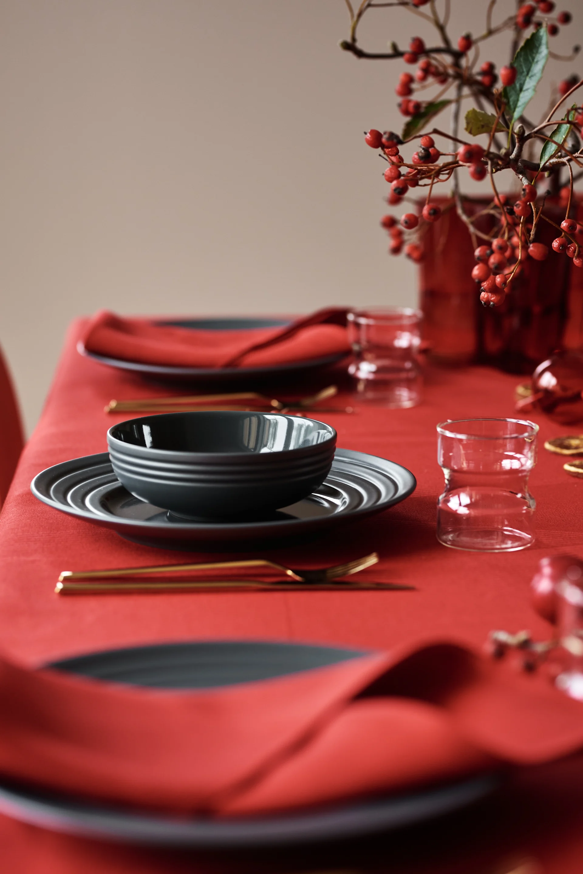 A festive table set with dark gray plates, gold cutlery, clear glasses, red napkins, and a vase of red berries on a red tablecloth.