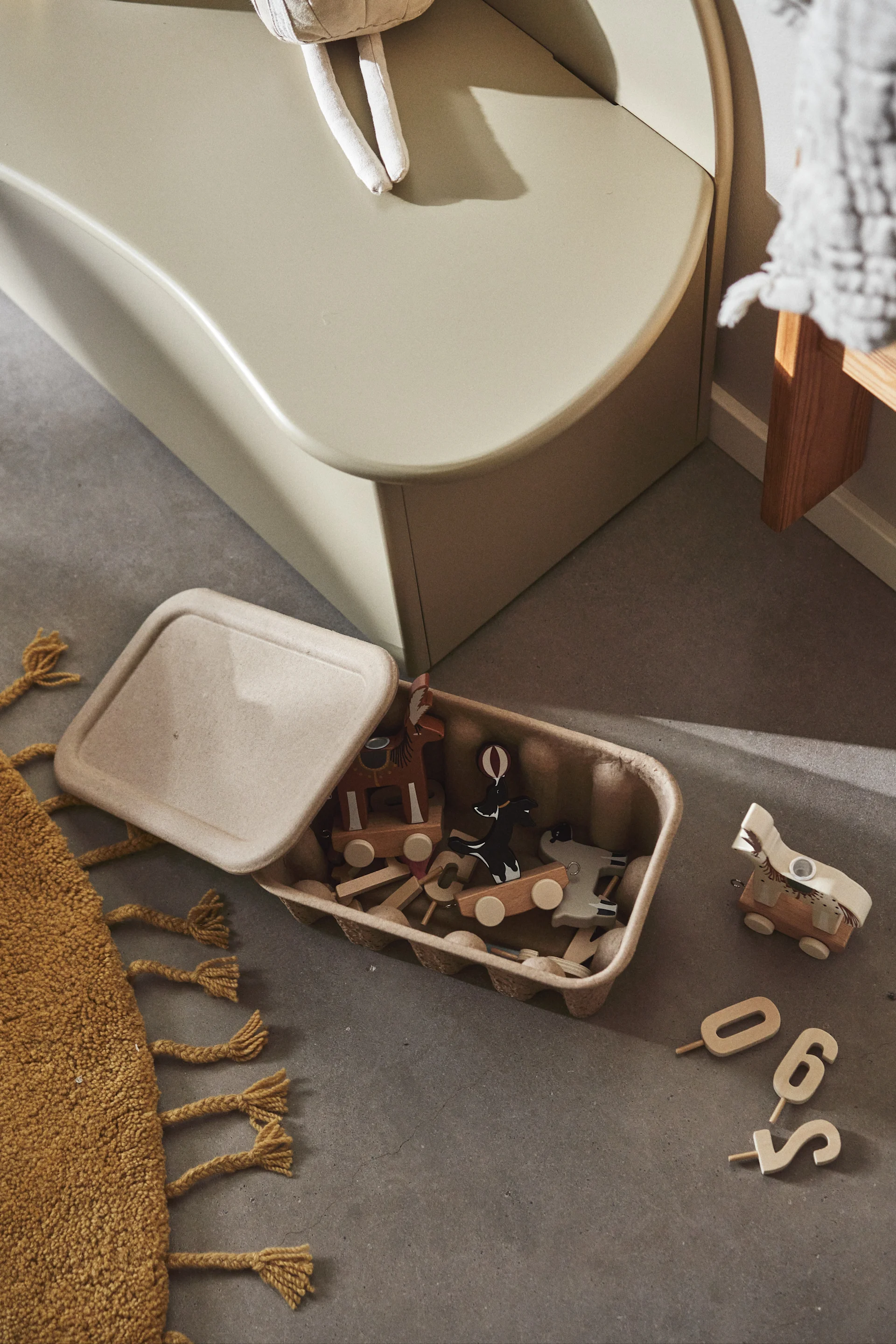 An open container of wooden toy animals and numbers on a concrete floor next to a yellow rug and a light green children's slide.