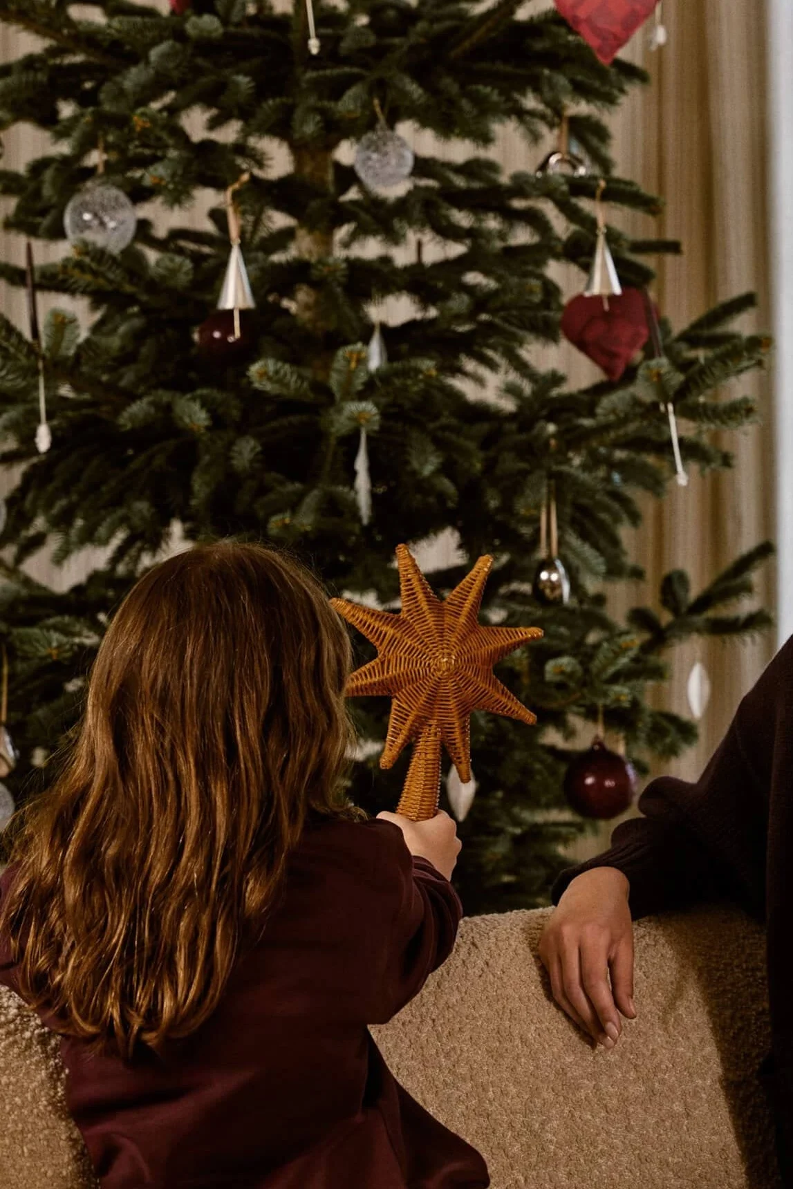 A child holds a woven star ornament in front of a decorated Christmas tree with red hearts and silver bells, next to an adult.
