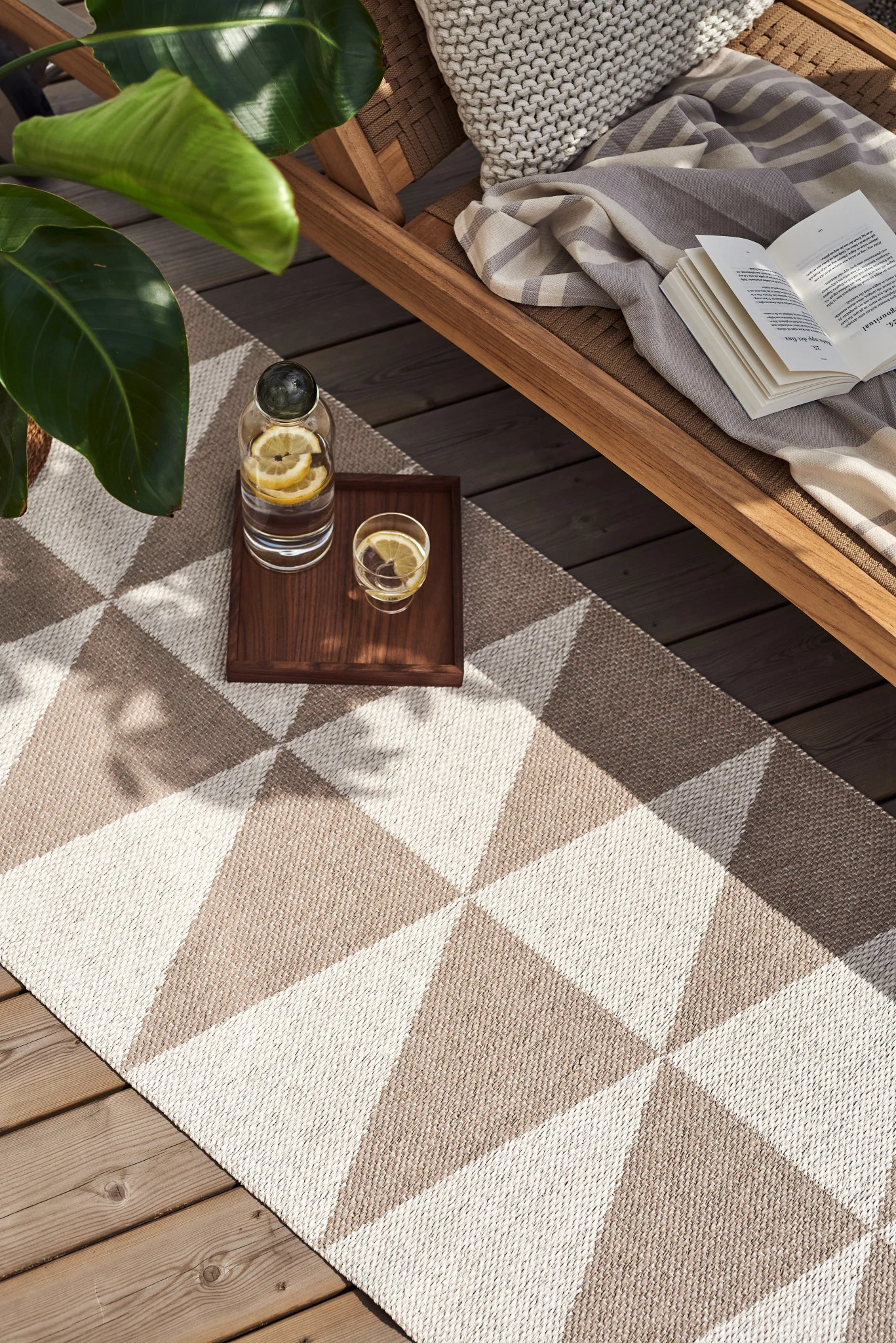 Outdoor lounge area with a geometric rug, wooden chair, open book, and lemon water on a tray.