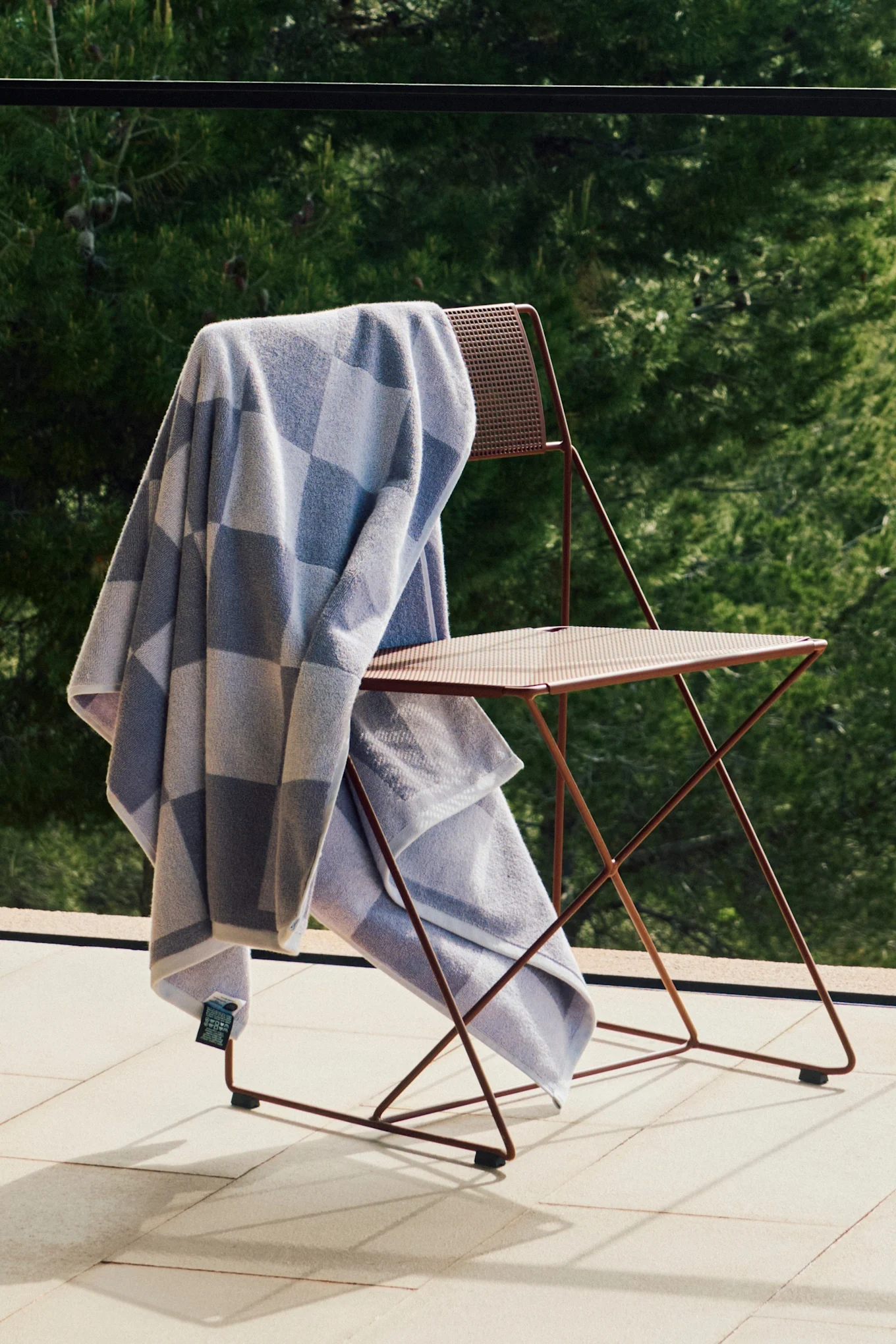 Light blue and grey checkered towel on a reddish-brown metal folding chair on a sunny patio with green foliage.