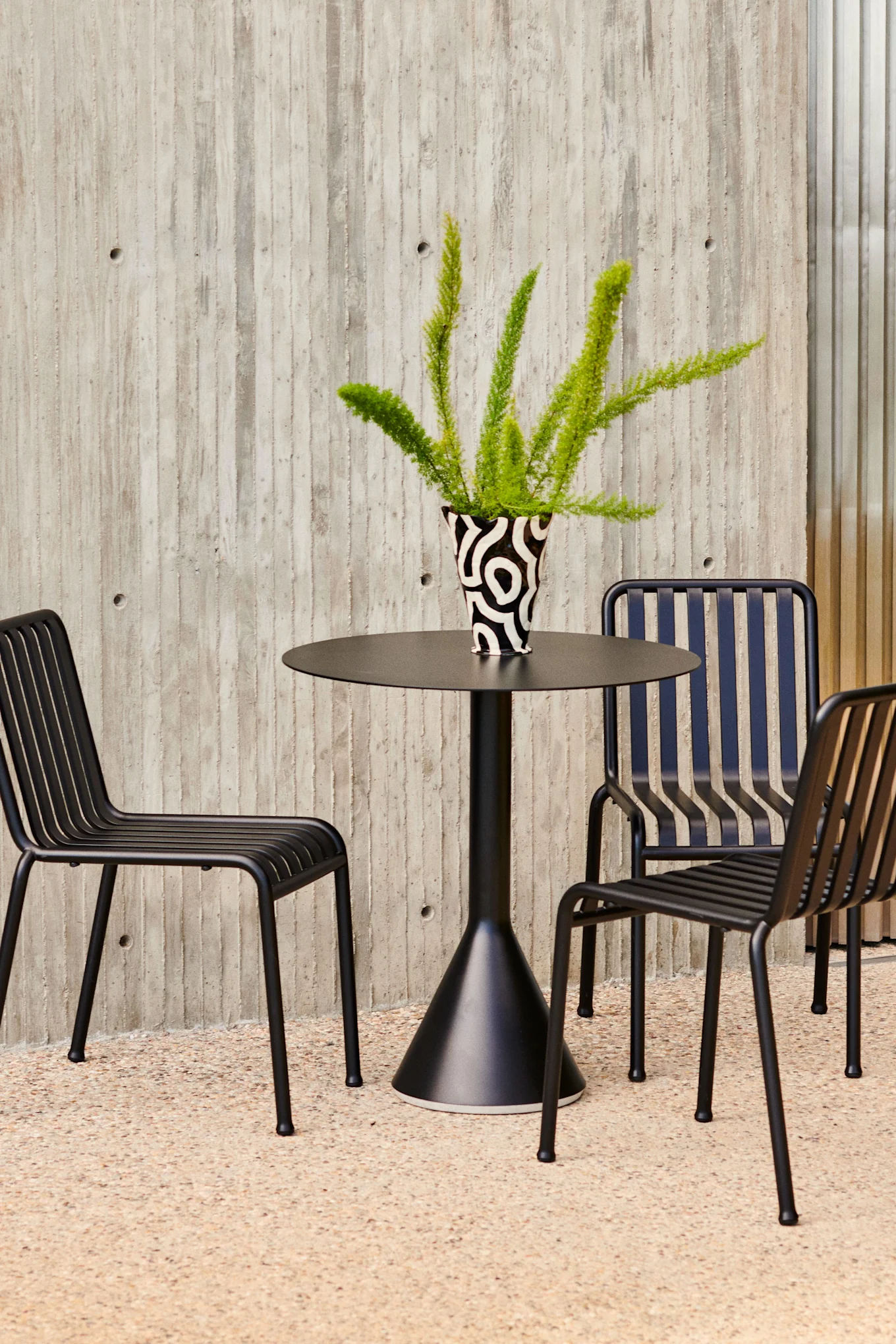A black round table with a patterned vase of green foliage and three black slatted chairs on a textured floor against a concrete wall.