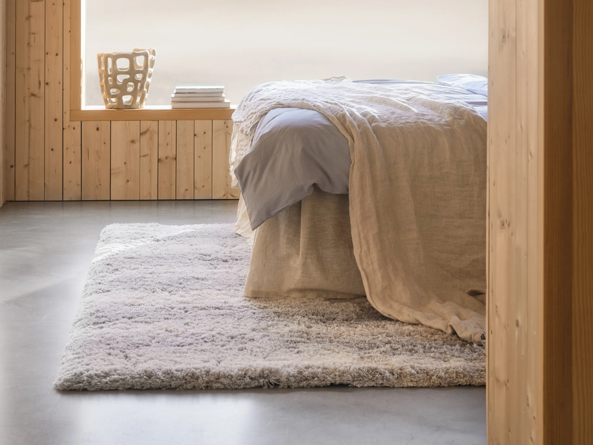 Cozy bedroom with a bed, light blue sheets, beige blanket, shaggy rug, wood paneled walls, and a decorative vase.