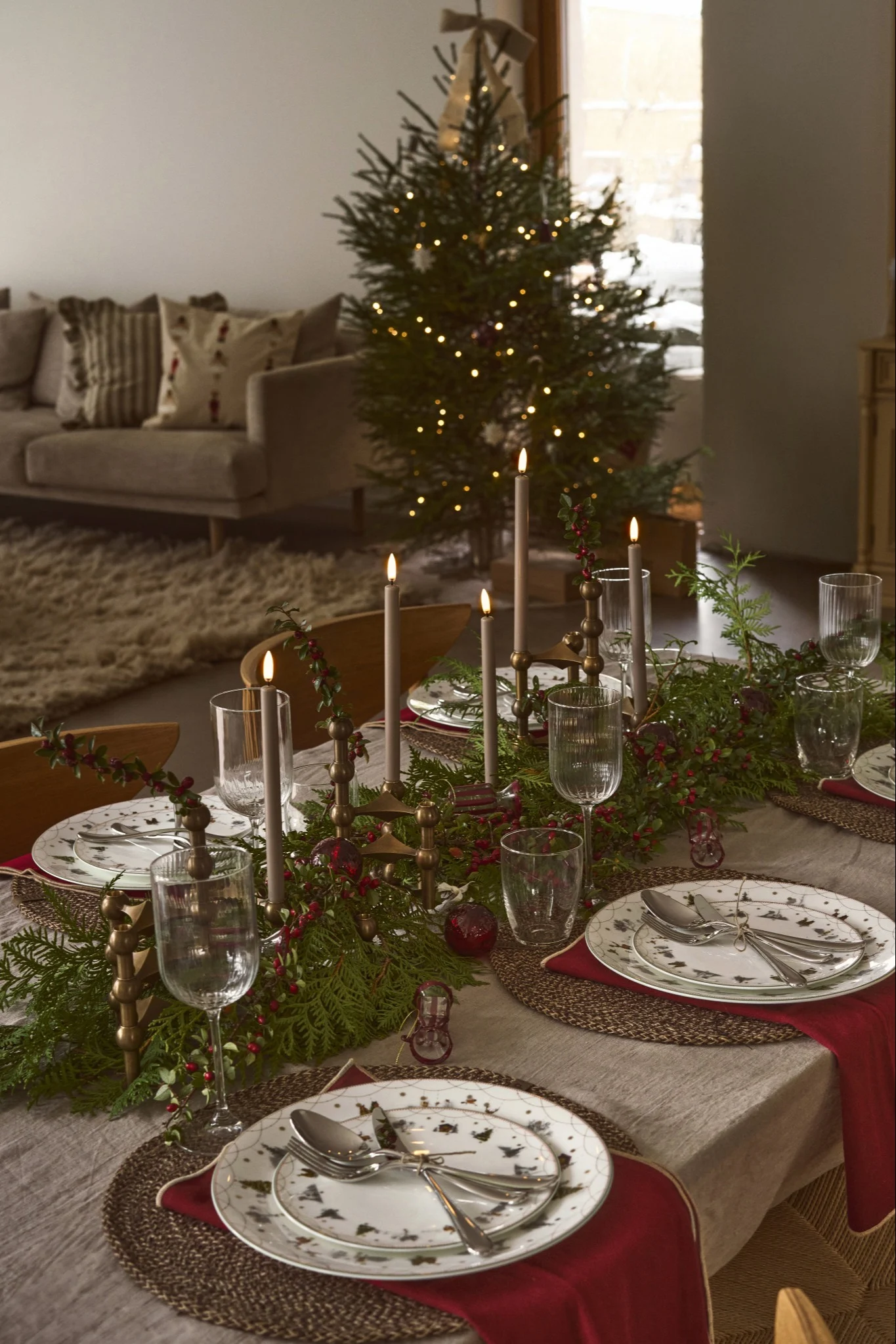 Christmas dining table set with lit candles, evergreen garland, red berries, patterned plates, and a decorated tree.