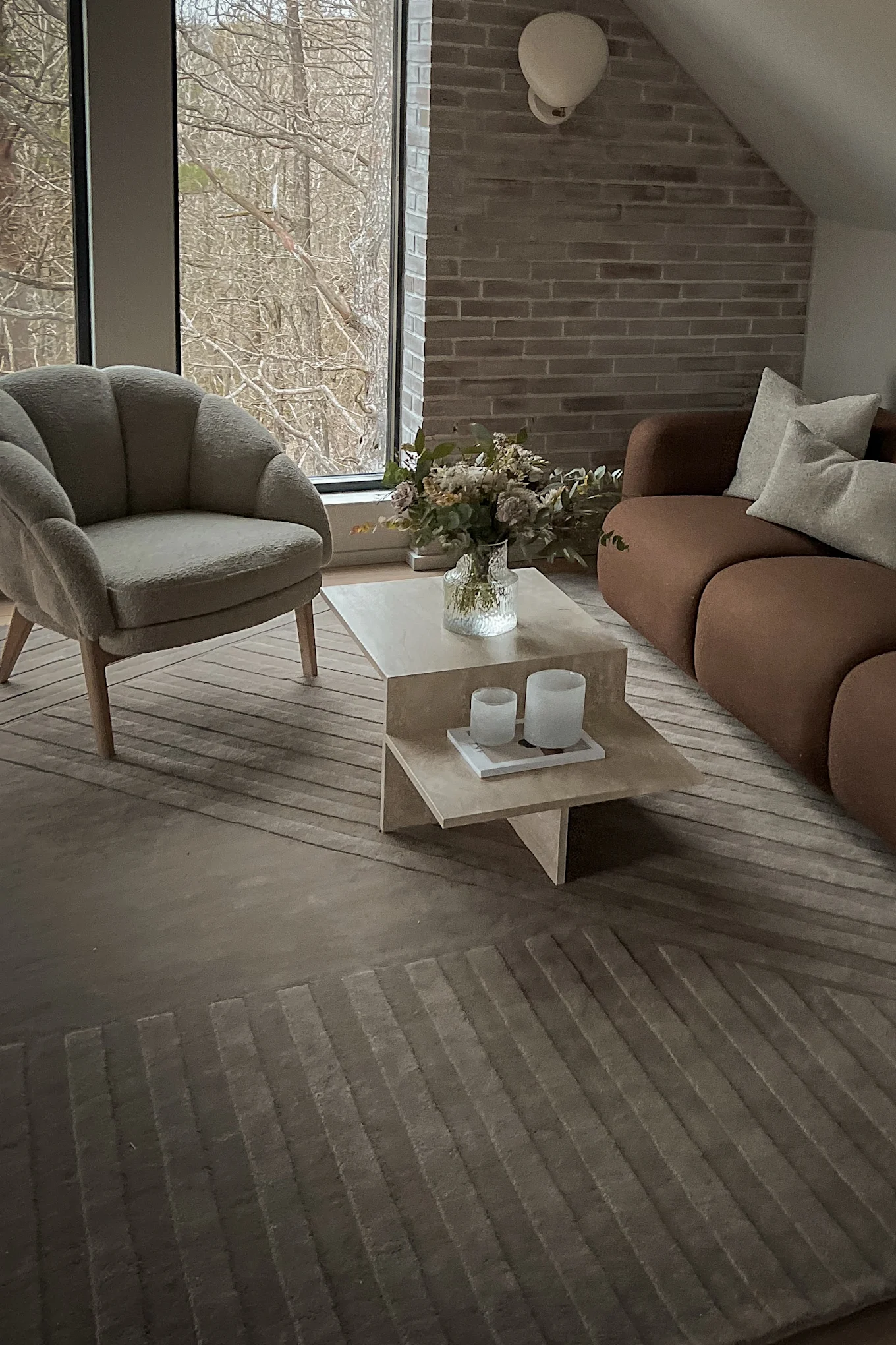A modern living room with a grey armchair, brown sofa, beige coffee table with flowers, and a window overlooking bare trees.