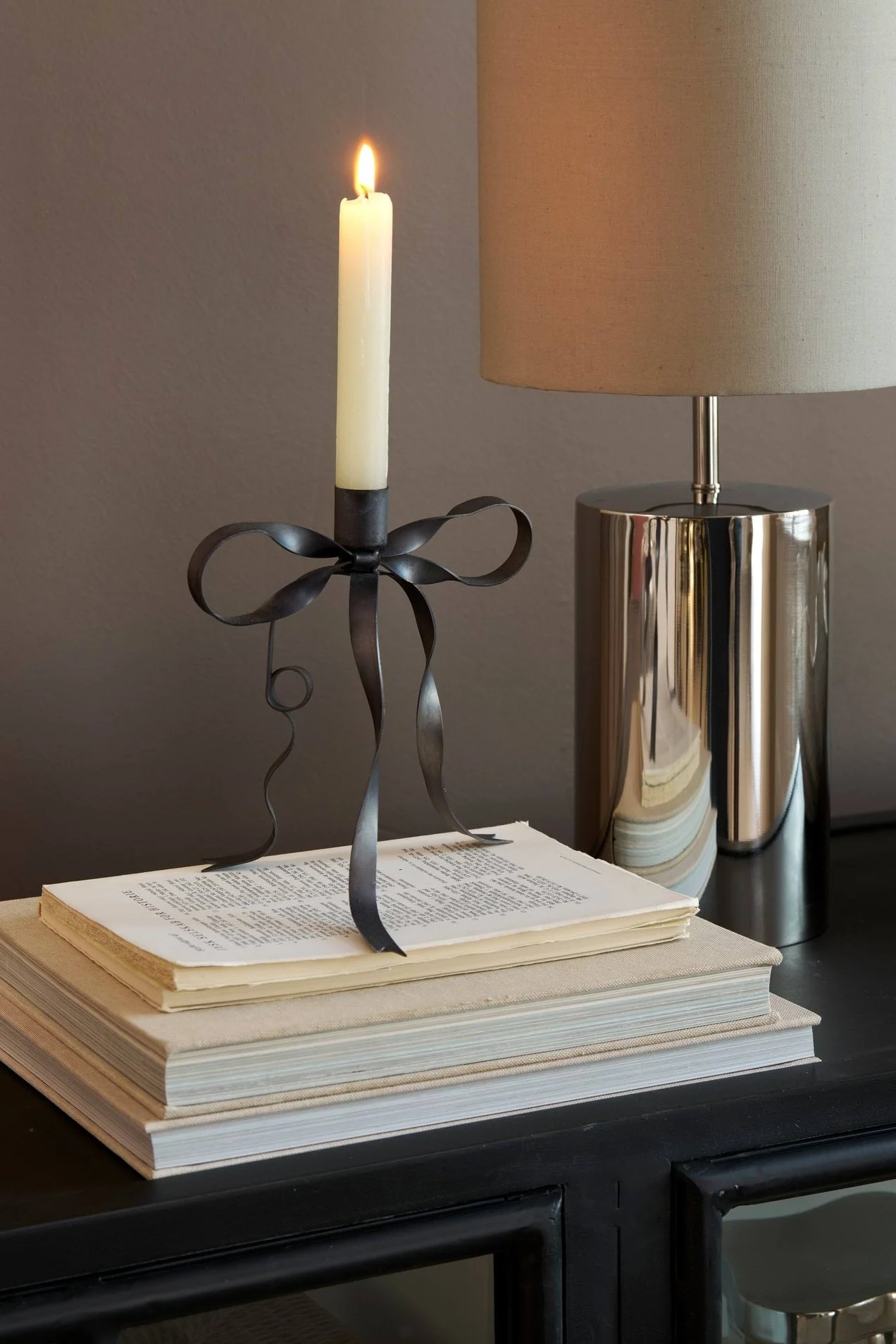 A lit white candle in a dark metal bow-shaped candlestick on a stack of books, next to a chrome table lamp with a fabric shade.