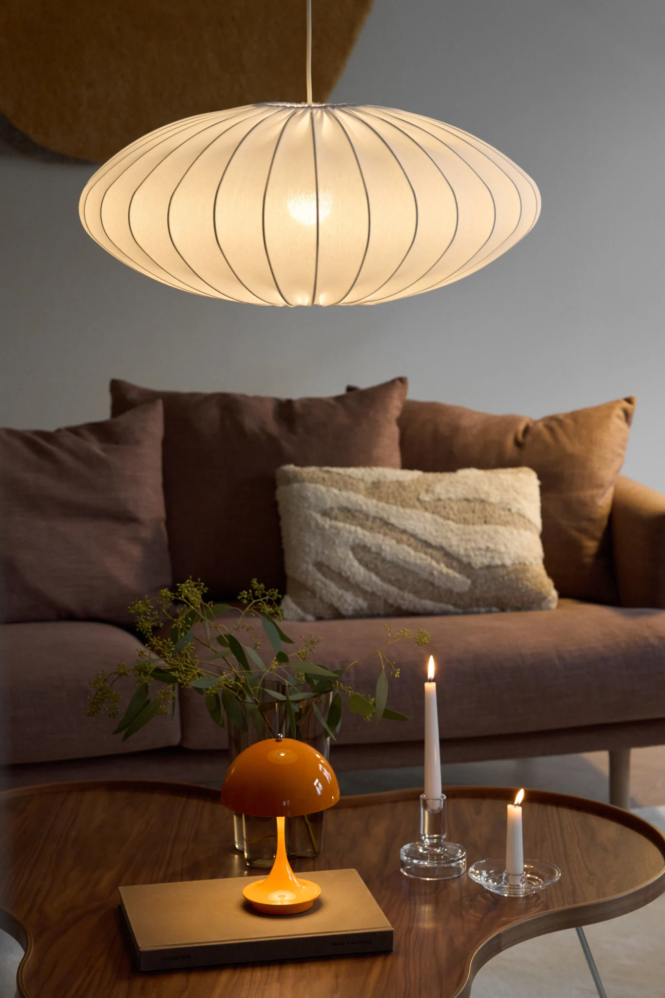 An oval, ribbed pendant light illuminates a living room with a brown sofa, a wooden coffee table, an orange mushroom lamp, and two lit candles.
