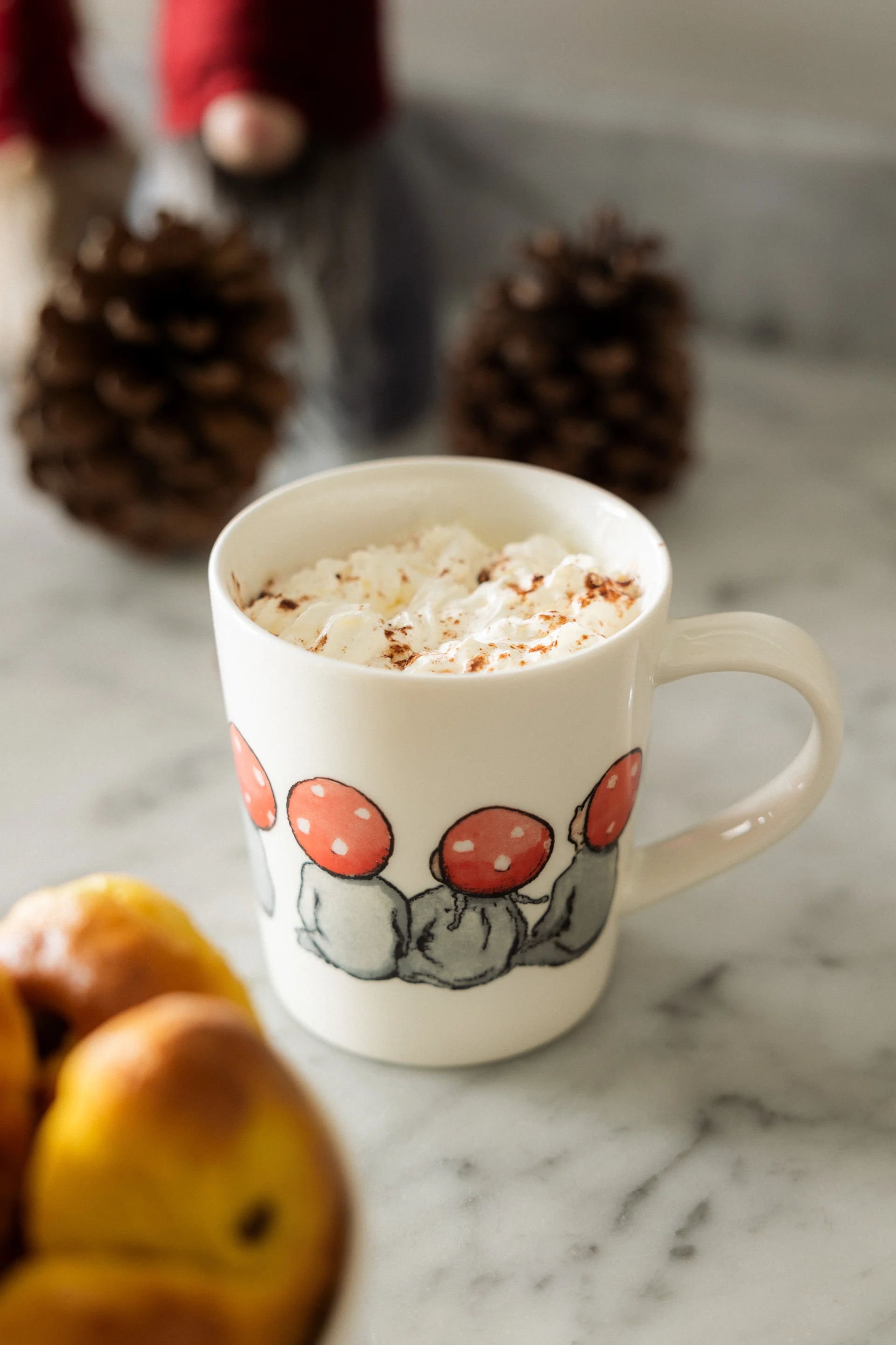 Mug with whipped cream and cinnamon, decorated with mushroom people, on a marble counter with pinecones and fruit.