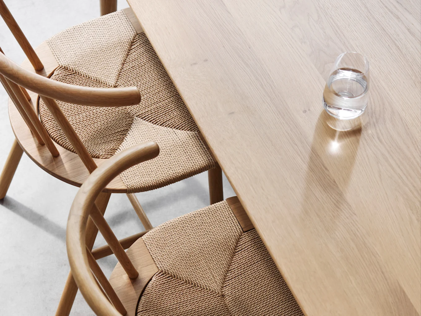 Overhead view of a light wood dining table and two chairs with woven seats, with a glass of water on the table.