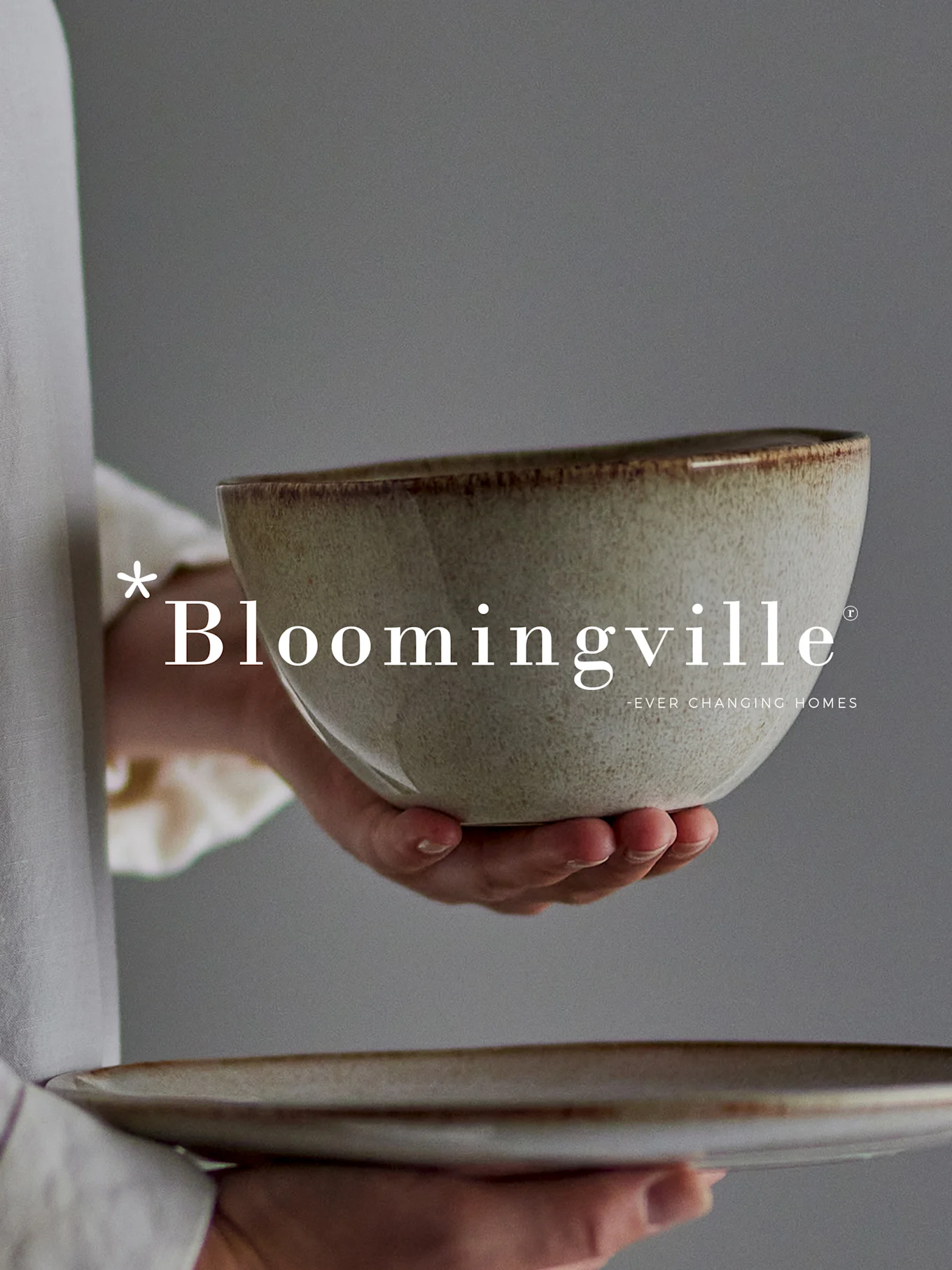Hands holding a rustic, speckled ceramic bowl and plate against a grey background. Bloomingville logo.