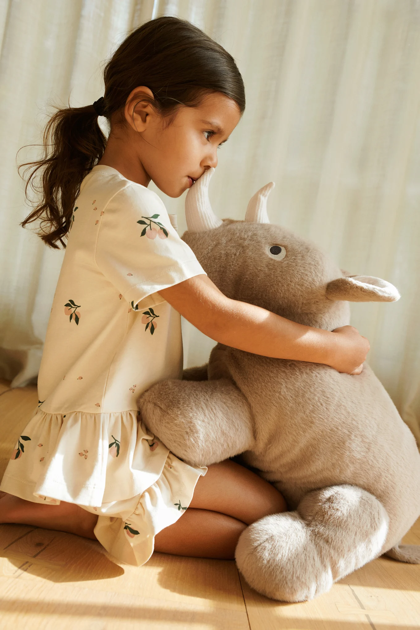 Young girl in patterned dress kneeling and hugging a large plush toy with horns.