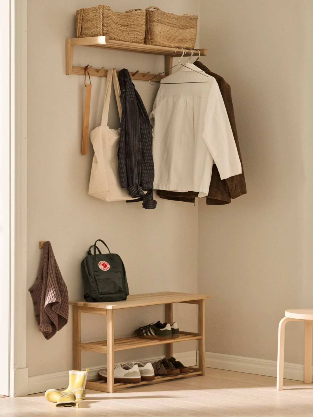 A wooden coat rack with woven baskets, hanging clothes, a green backpack, a shoe rack with sneakers and sandals, and yellow rain boots in an entryway.