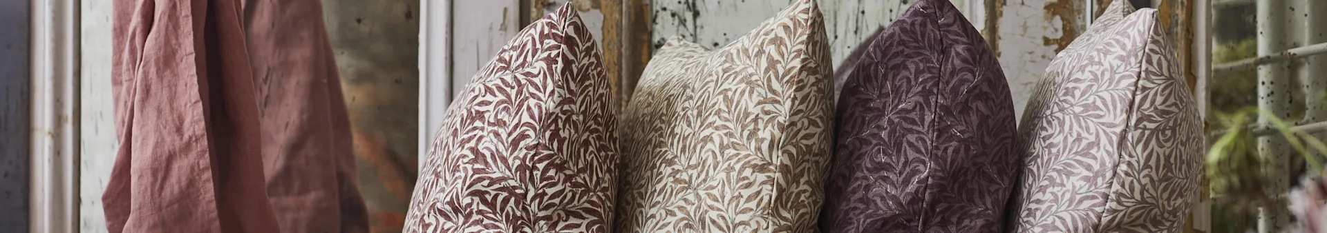 Four patterned pillows in shades of brown and purple, with a pink fabric draped on the left, against a rustic background.