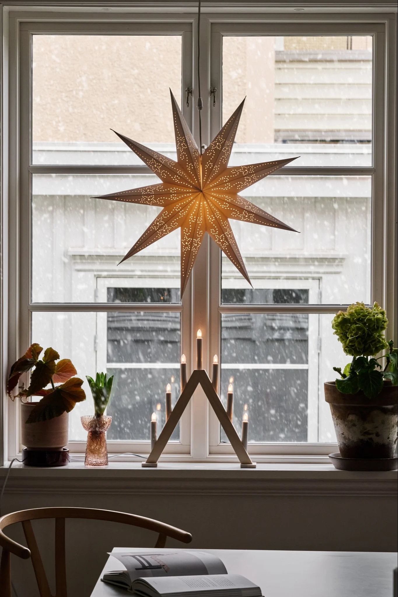 A pointed Advent candle holder stands in a large window alongside a hanging Christmas star and festive plants.
