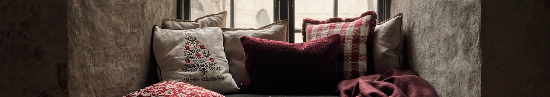 Cozy window seat with cushions: an embroidered Christmas tree cushion, a red textured, a red checkered, and a red throw.