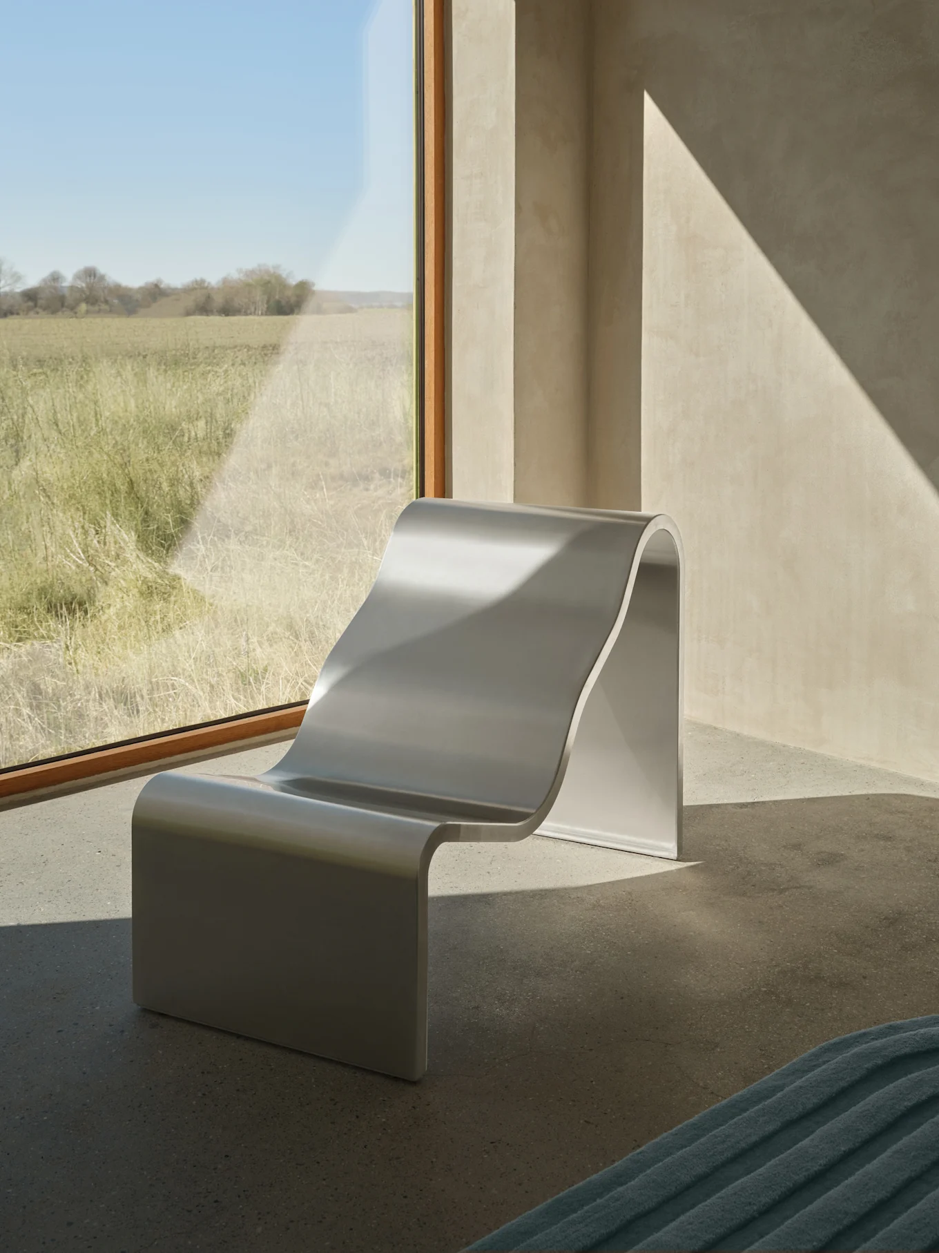 A modern silver S-shaped lounge chair sits by a large window overlooking a sunny grassy field, with diagonal sunlight patterns on the walls.