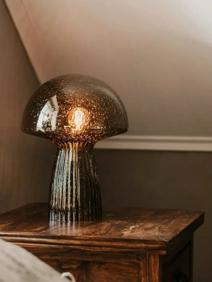 A mushroom-shaped table lamp with a textured, smoked glass shade and ribbed base, glowing warmly on a wooden nightstand.