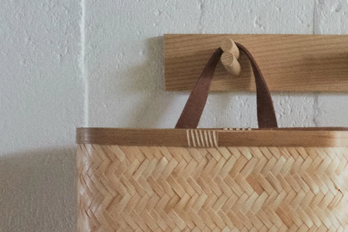 Woven basket with brown leather handle hanging on a wooden hook against a white wall.