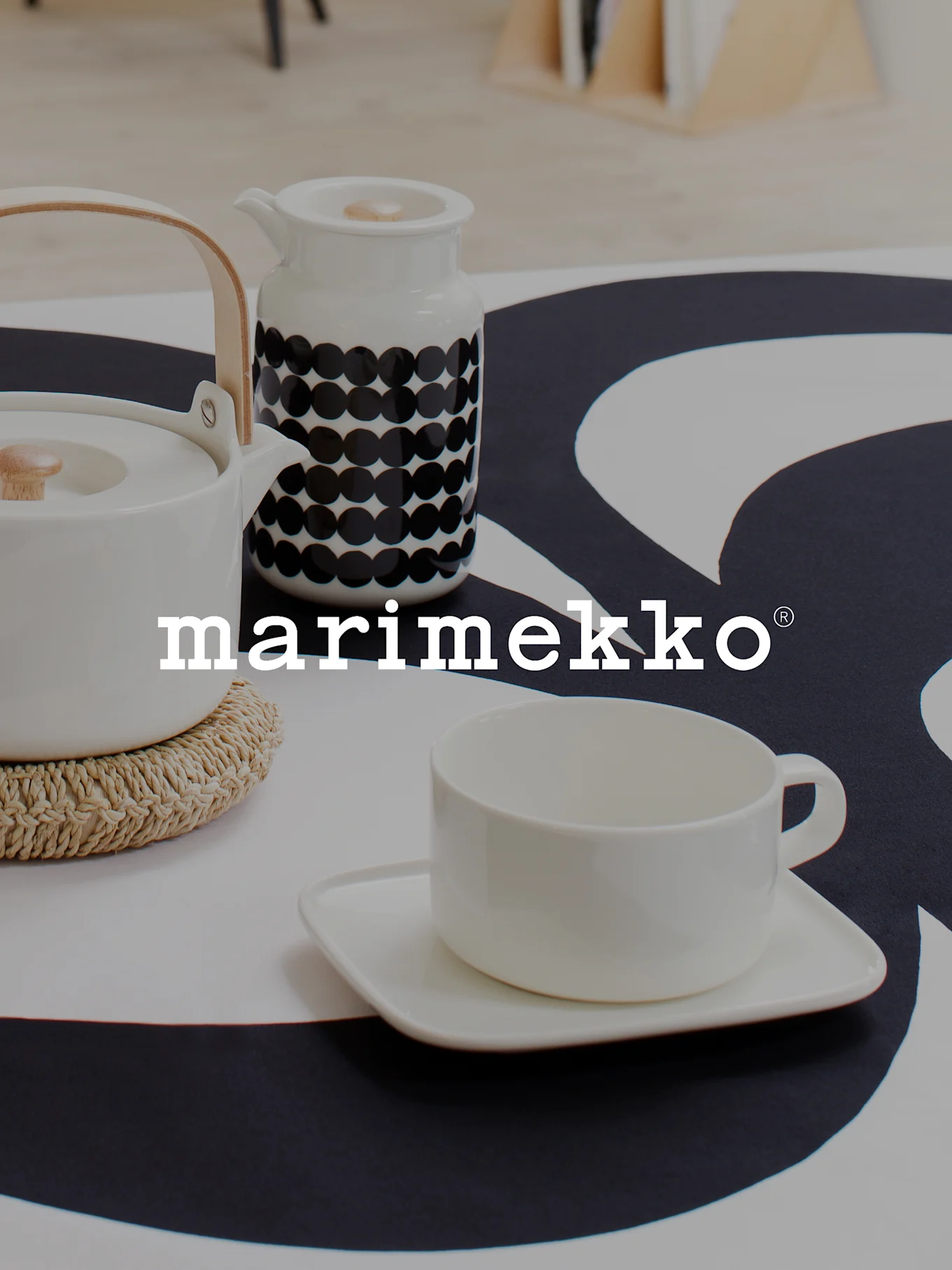 Marimekko logo over a white teapot with wooden handle, a white polka dot creamer, and a white teacup on a black and white patterned tablecloth.