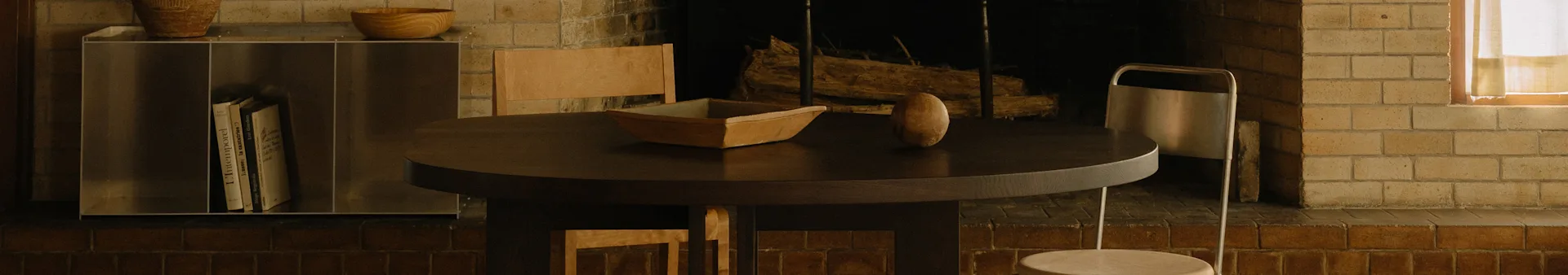 A cozy dining area with a round dark wood table, a wooden bowl, an oval object, chairs, and a brick fireplace.