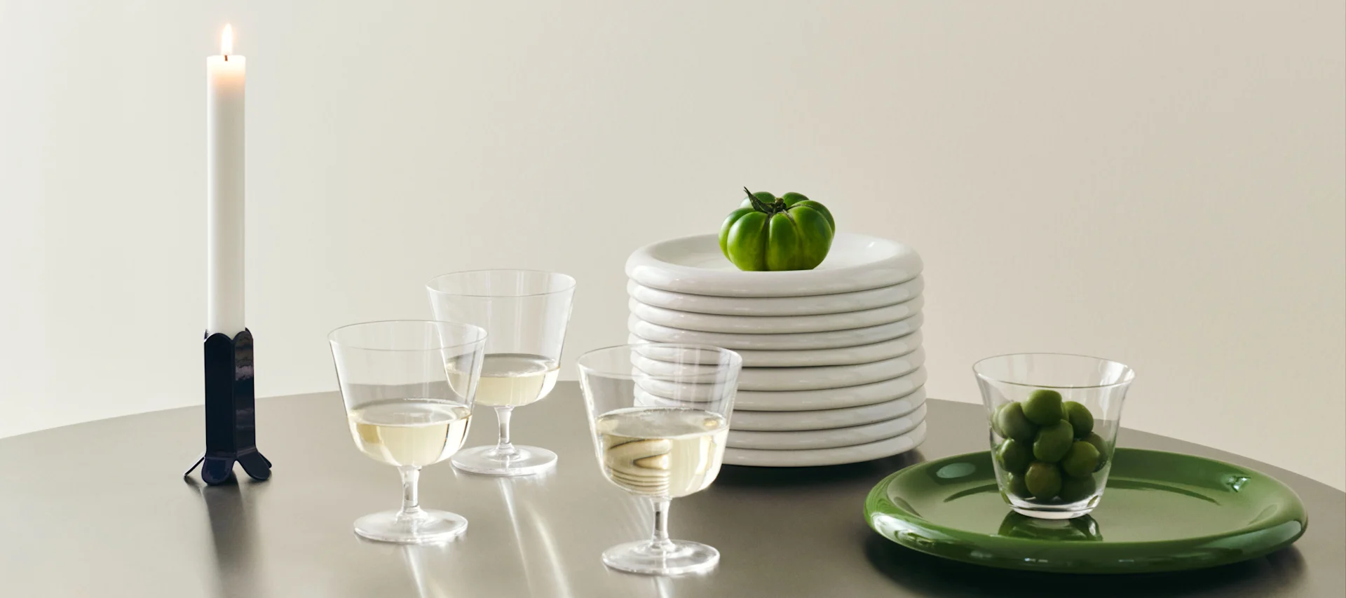 Table setting with candle, wine glasses, stacked white plates with a green tomato, and green olives on a green plate.