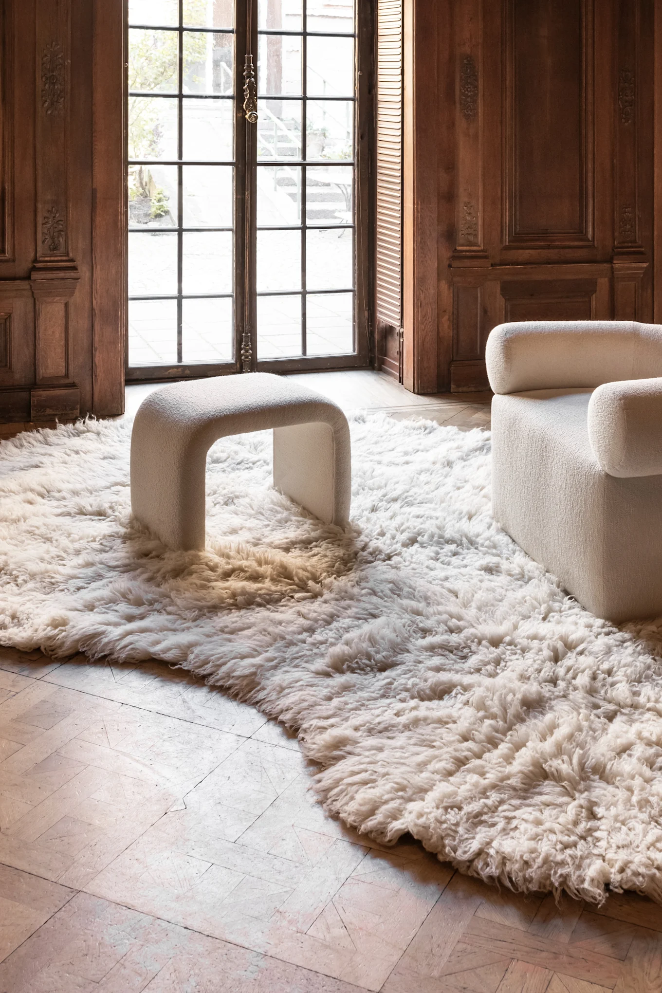 A cozy room with dark wood paneling, featuring a fluffy white rug, a U-shaped white stool, and part of a white armchair by a large window.