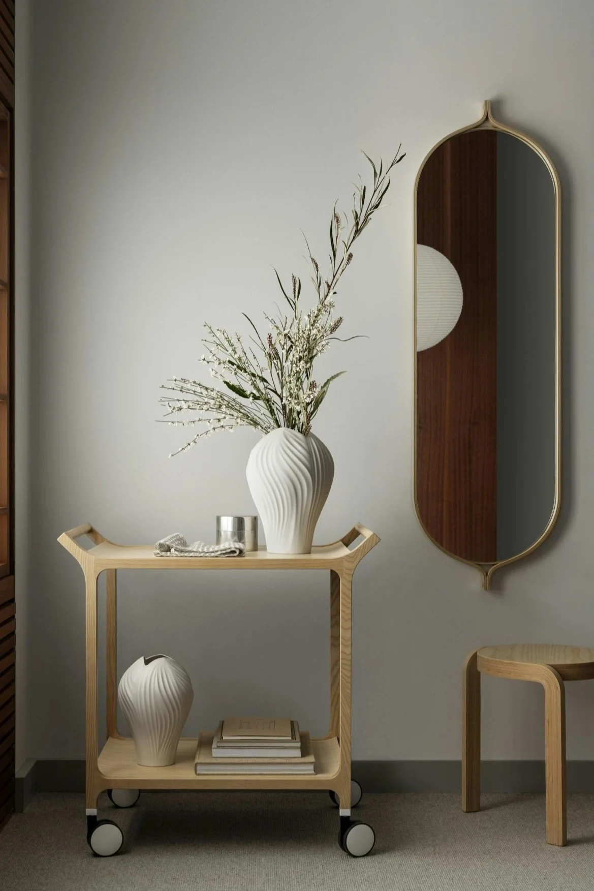 A light wood rolling cart with white vases and flowers, next to a tall, arched mirror reflecting a dark wood panel.