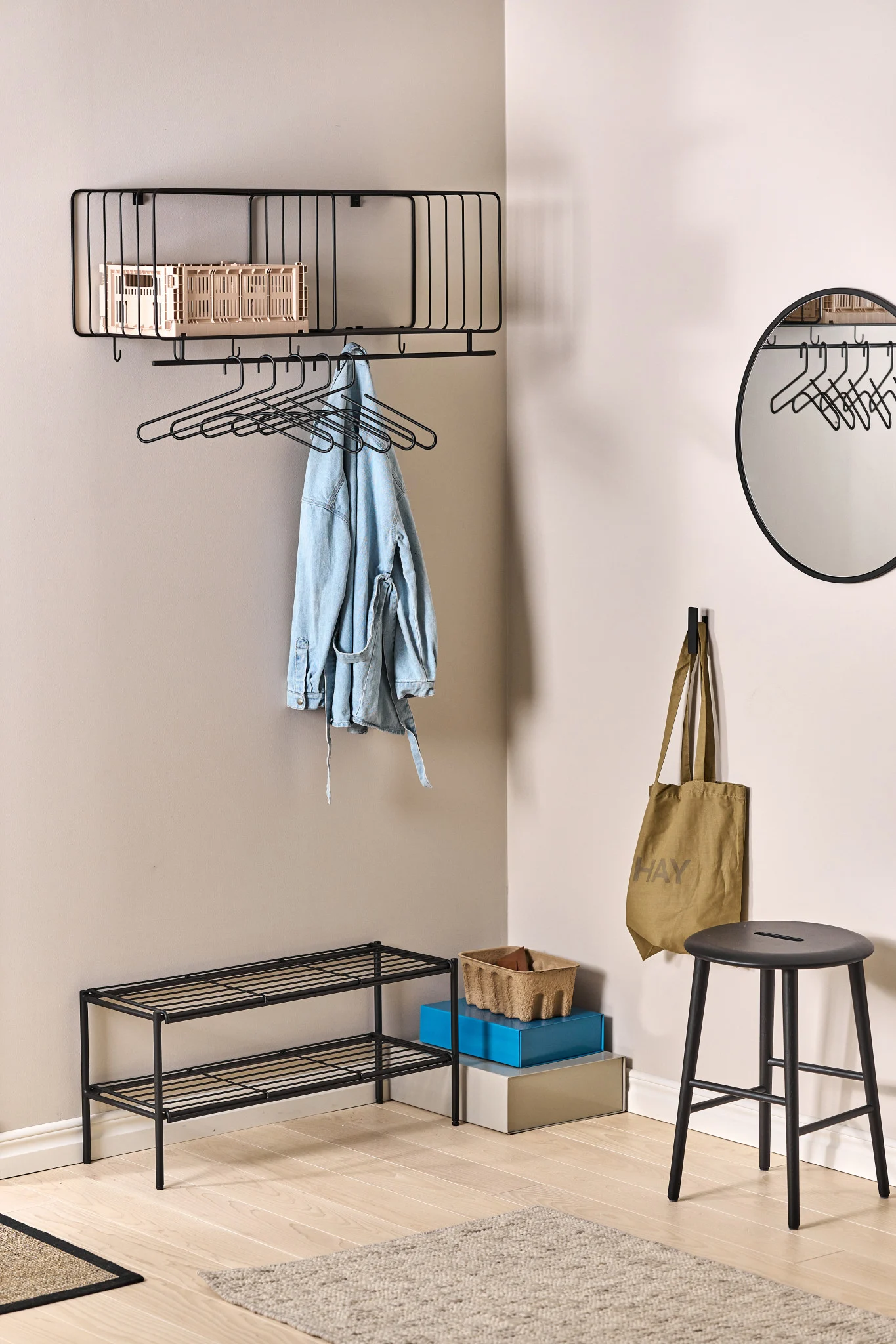 A modern entryway with a black wall shelf, denim jacket on hangers, a shoe rack, round mirror, and a black stool.