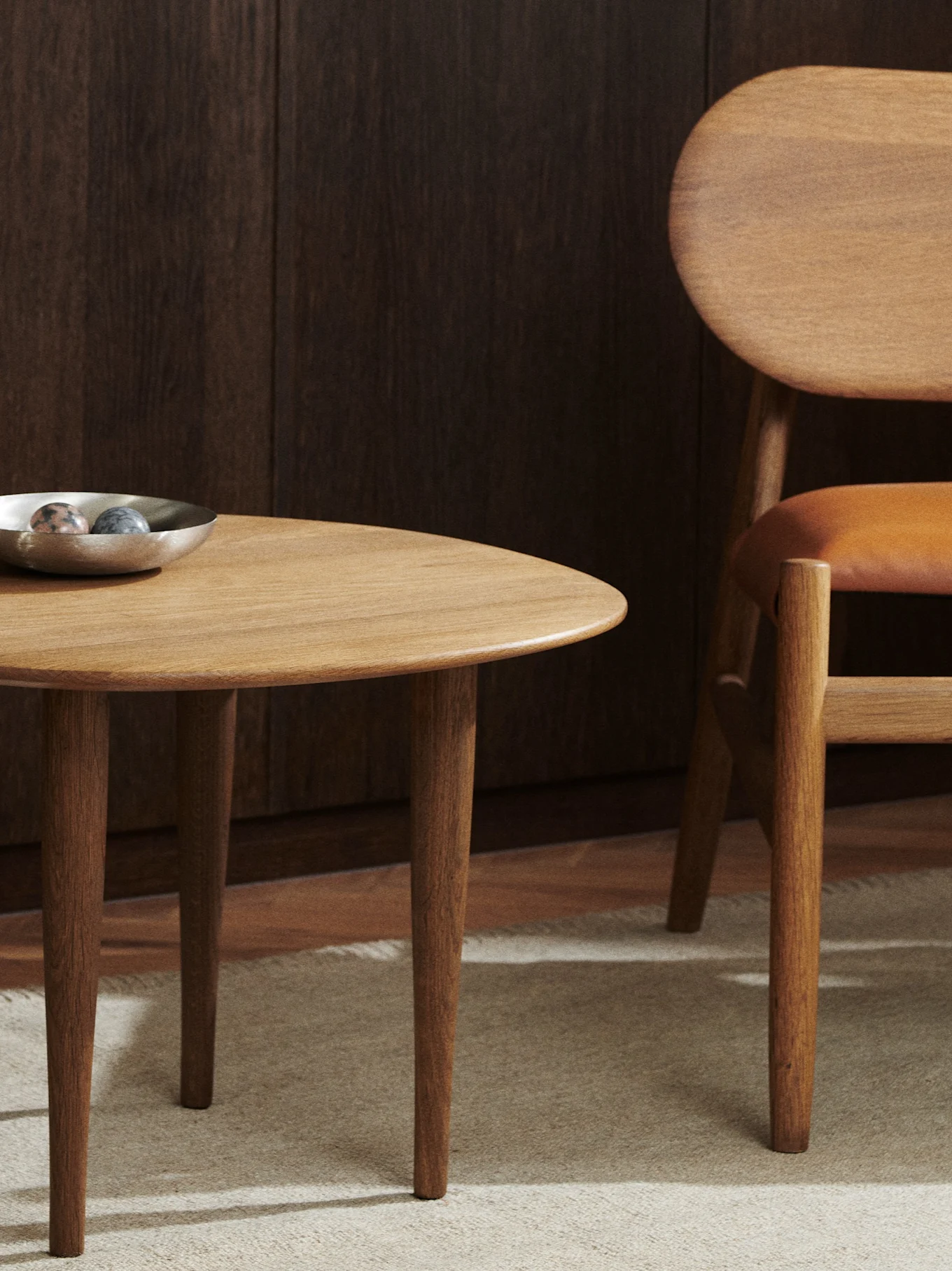 A round wooden table with a silver bowl and a wooden chair with a leather seat on a light rug.