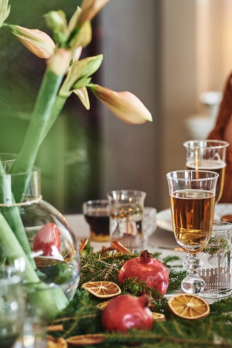 Festive table setting with amaryllis flowers, pomegranates, dried oranges, and various drinks.