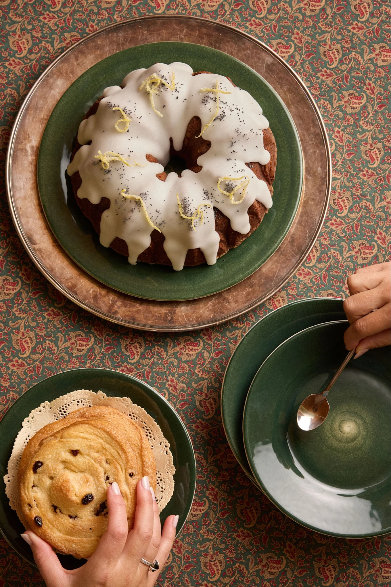 Overhead: lemon poppy seed Bundt cake, raisin pastry, and hands on a patterned tablecloth.