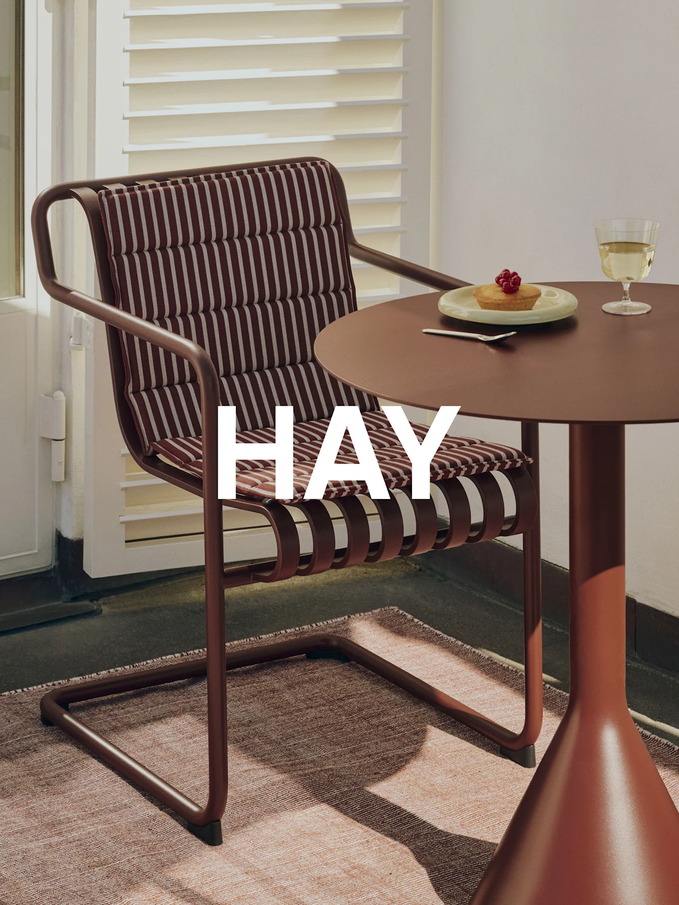 Outdoor setting with brown metal chair, striped cushion, matching table, tart, wine glass, and 'HAY' branding.
