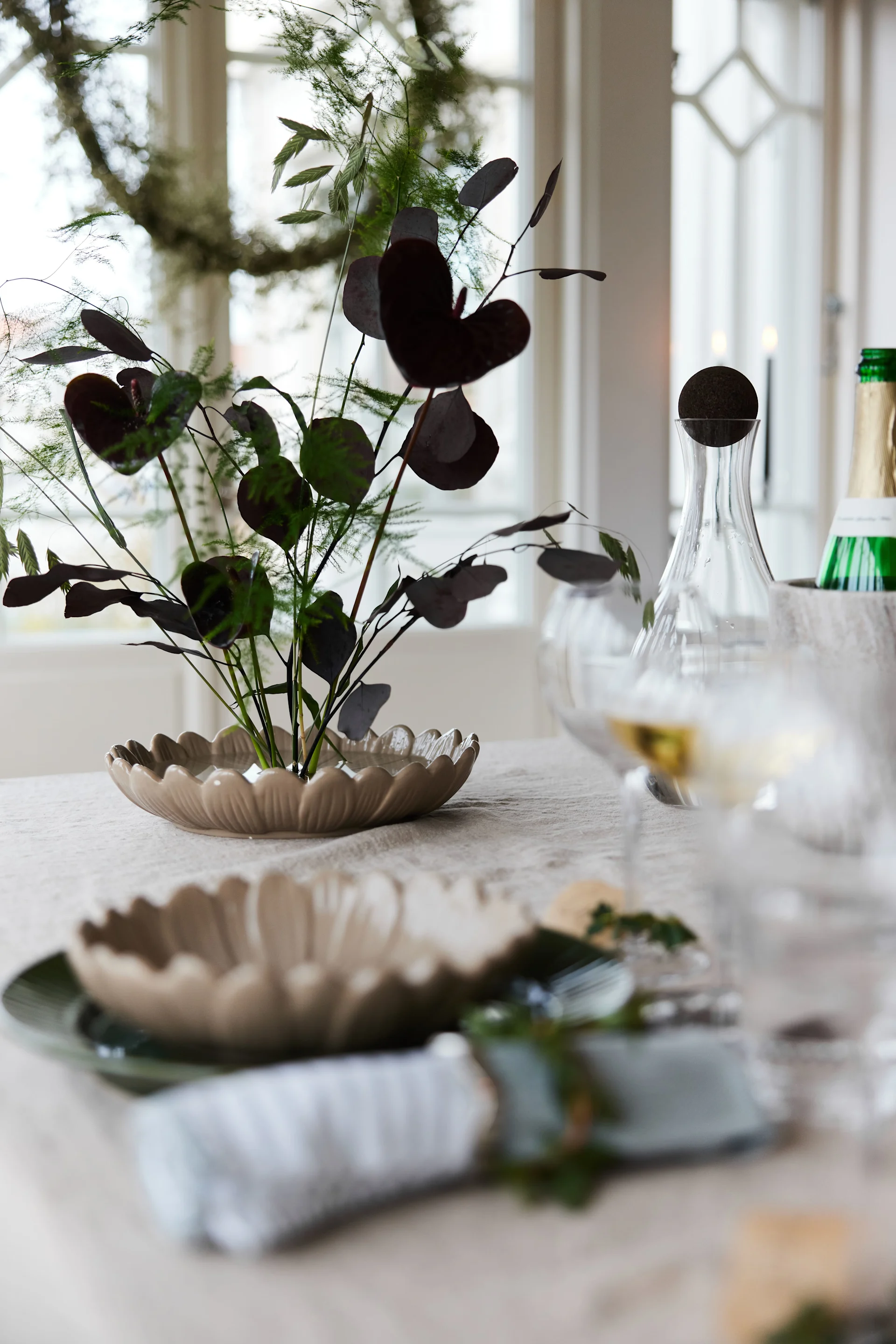 A festive table setting with a plant centerpiece in a scallop-edged bowl, a place setting with a green plate, and wine glasses with a carafe and bottle.