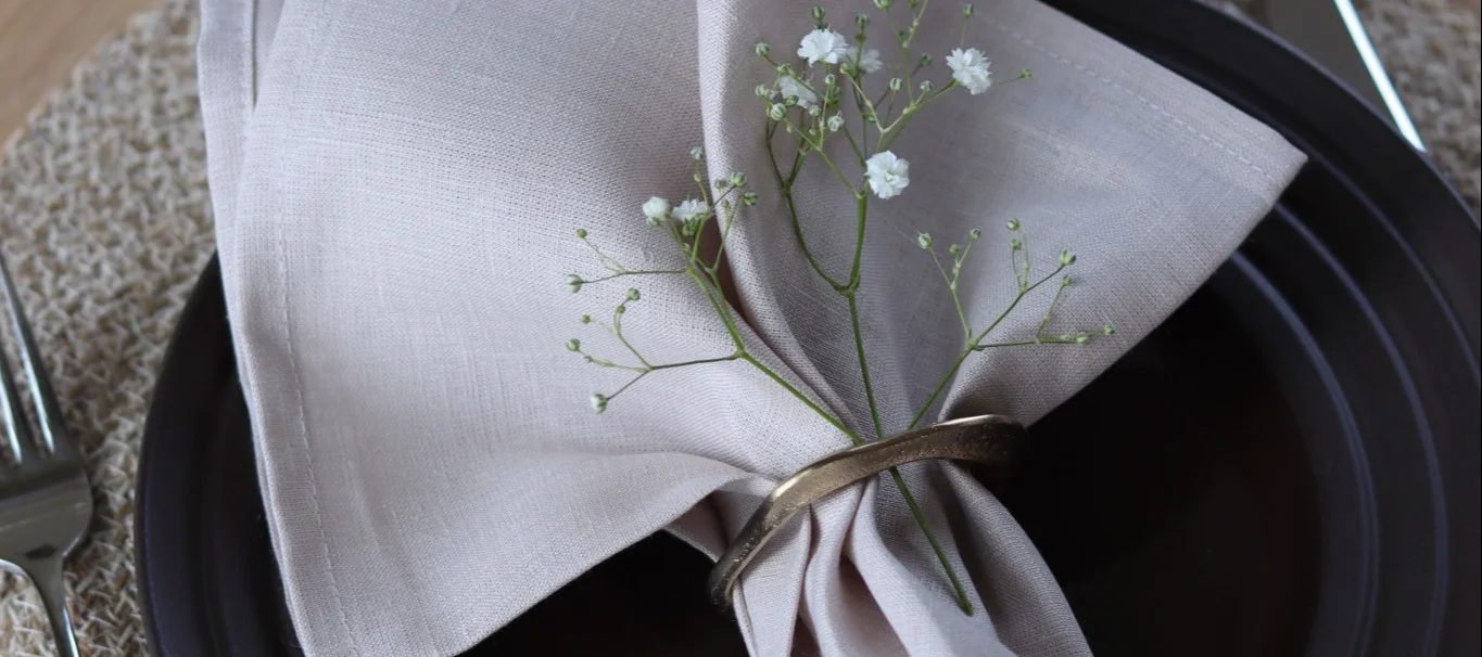 A light beige linen napkin with a gold napkin ring and baby's breath flowers on a dark plate.