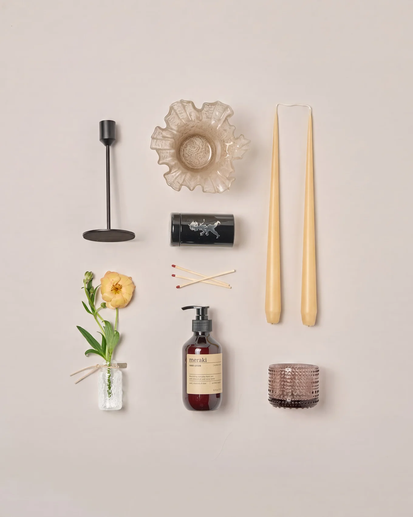 A flat lay of various home decor items: candlestick, ruffled bowl, tapered candles, matches, lotion, votive, and a ranunculus flower.