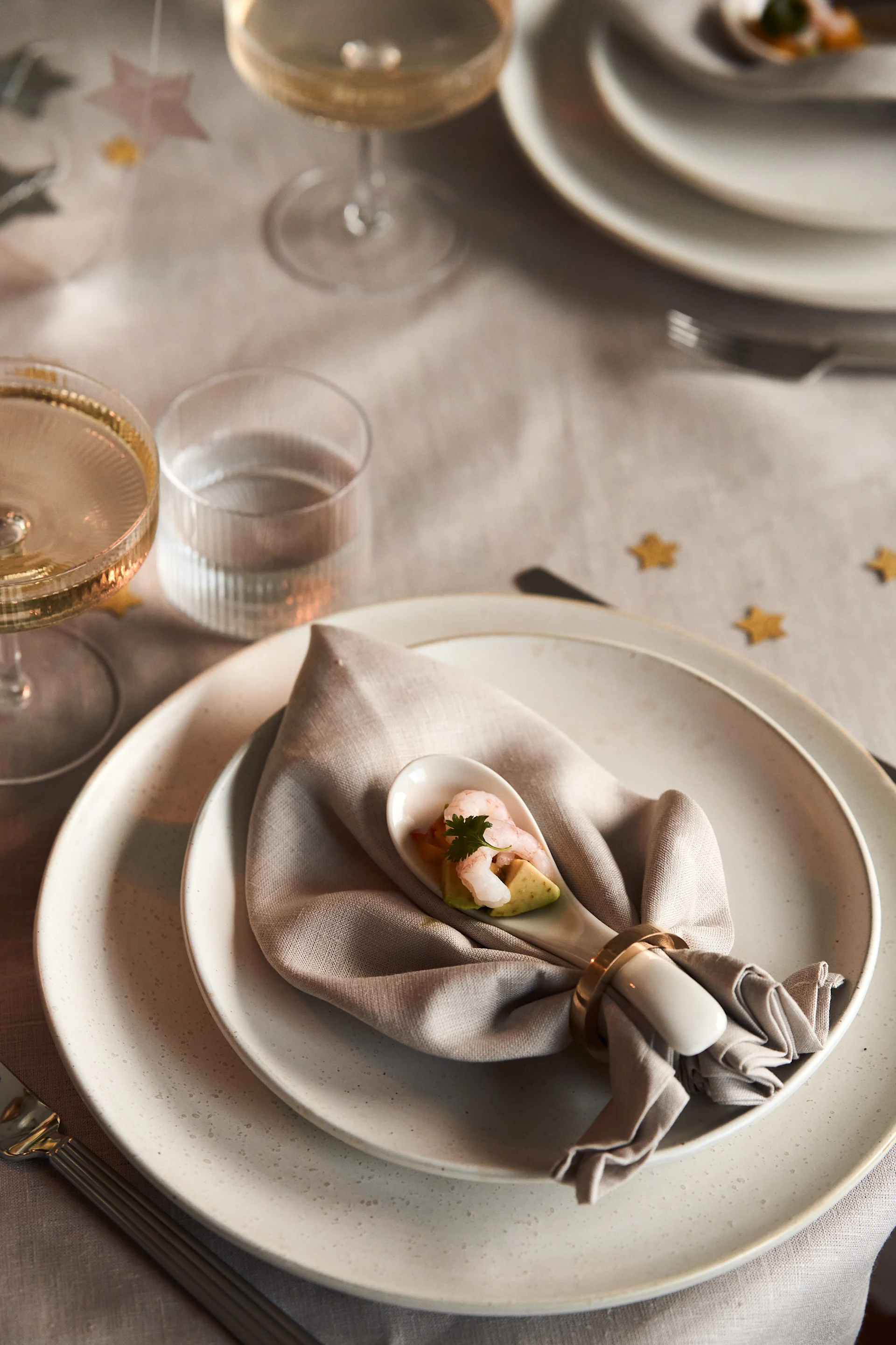 Elegant place setting with a plate, linen napkin, and spoon holding shrimp and avocado appetizer. Glasses and star confetti on the table.