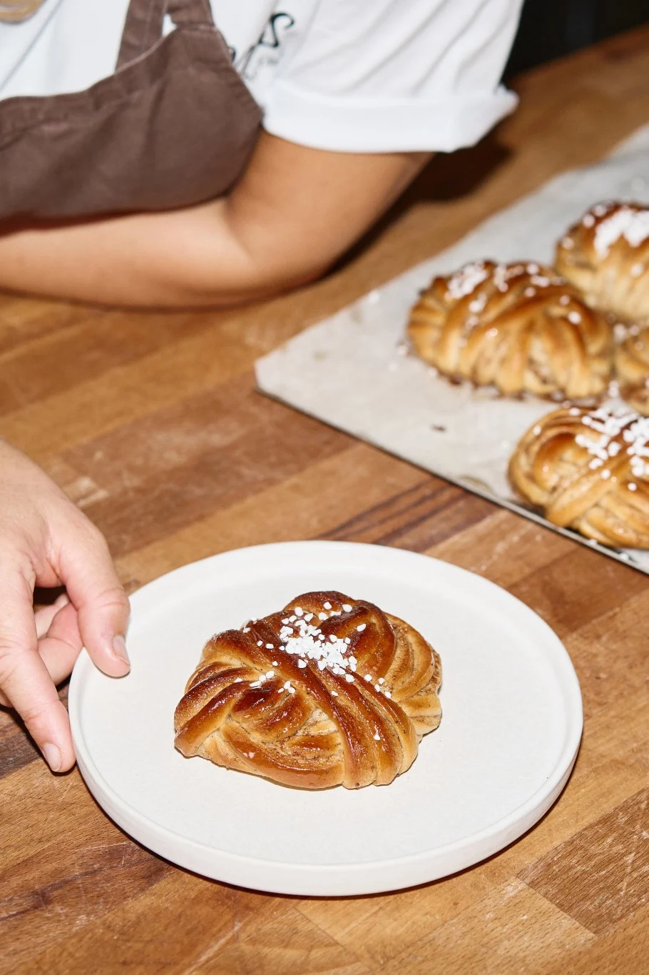 Hand places a sugar-sprinkled, braided cinnamon bun on a plate; more buns on a tray.