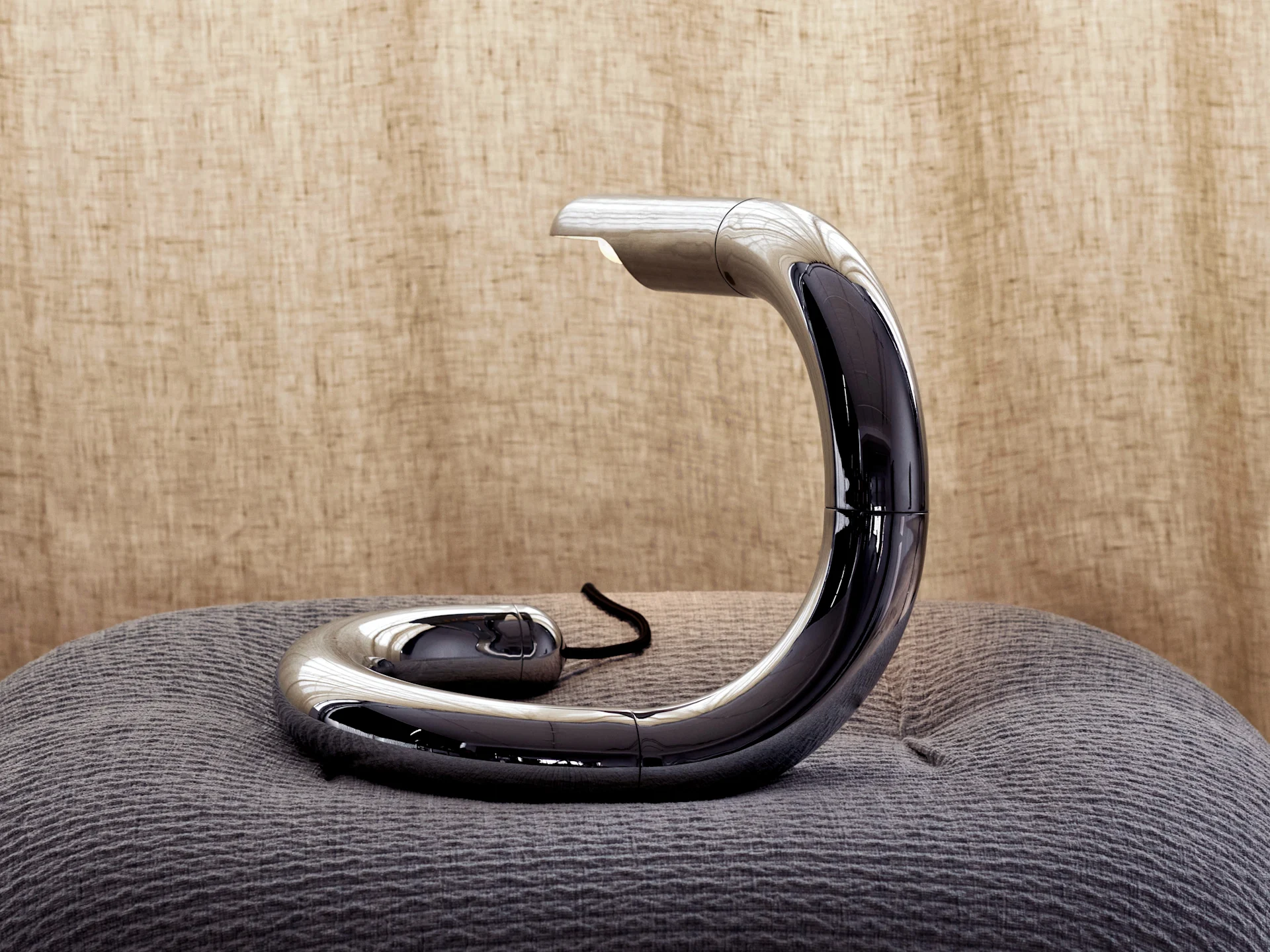 Sleek, C-shaped silver and black desk lamp on a textured gray cushion against a light brown background.