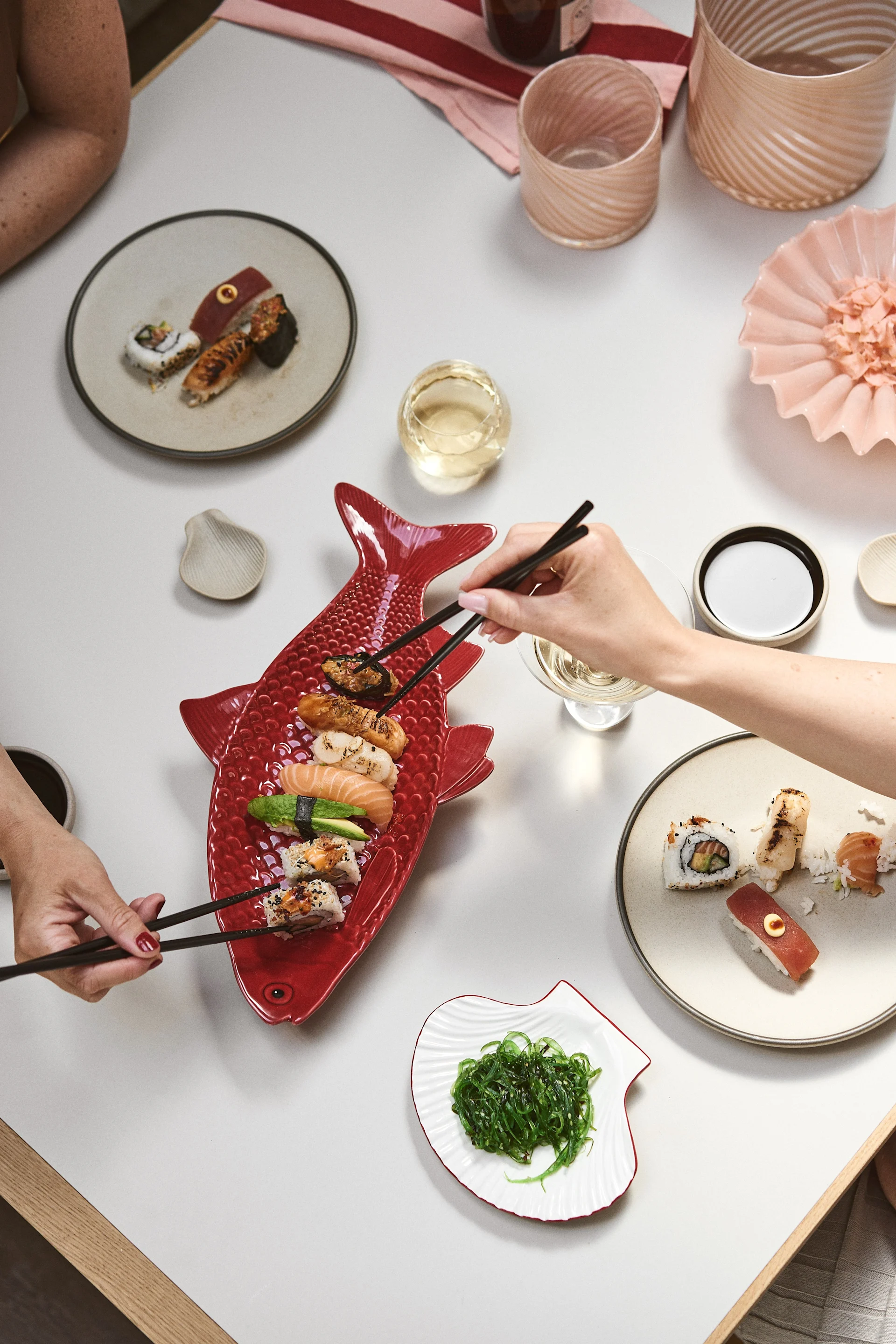 Two people using chopsticks to eat sushi from a red fish-shaped platter and a round plate on a light table.