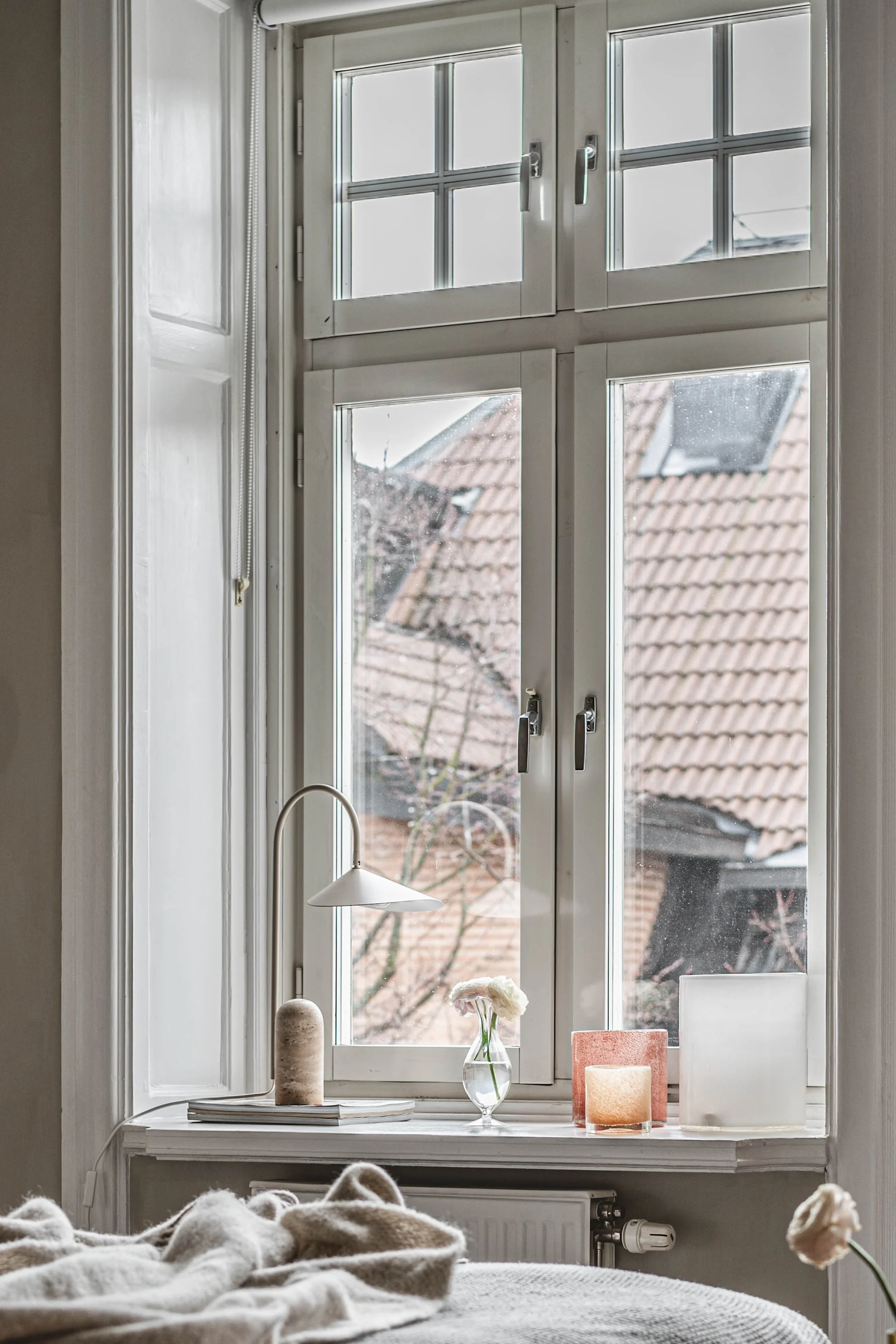 Window sill with a modern lamp, a white flower in a vase, and two decorative glass objects, overlooking a street with tiled roofs.