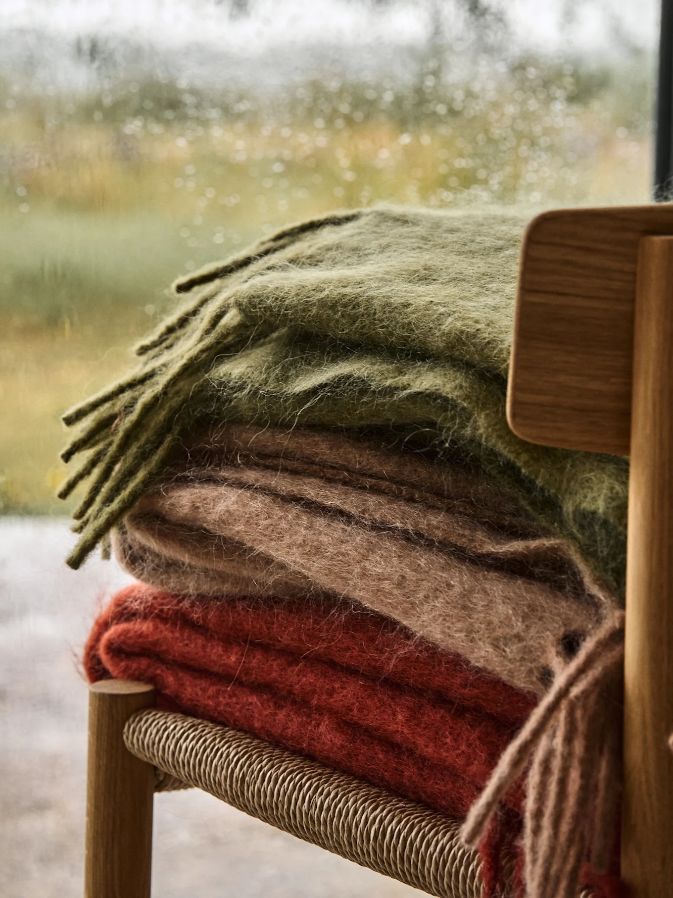 Stack of three folded blankets in olive green, light brown, and rust red on a wooden chair by a rainy window.