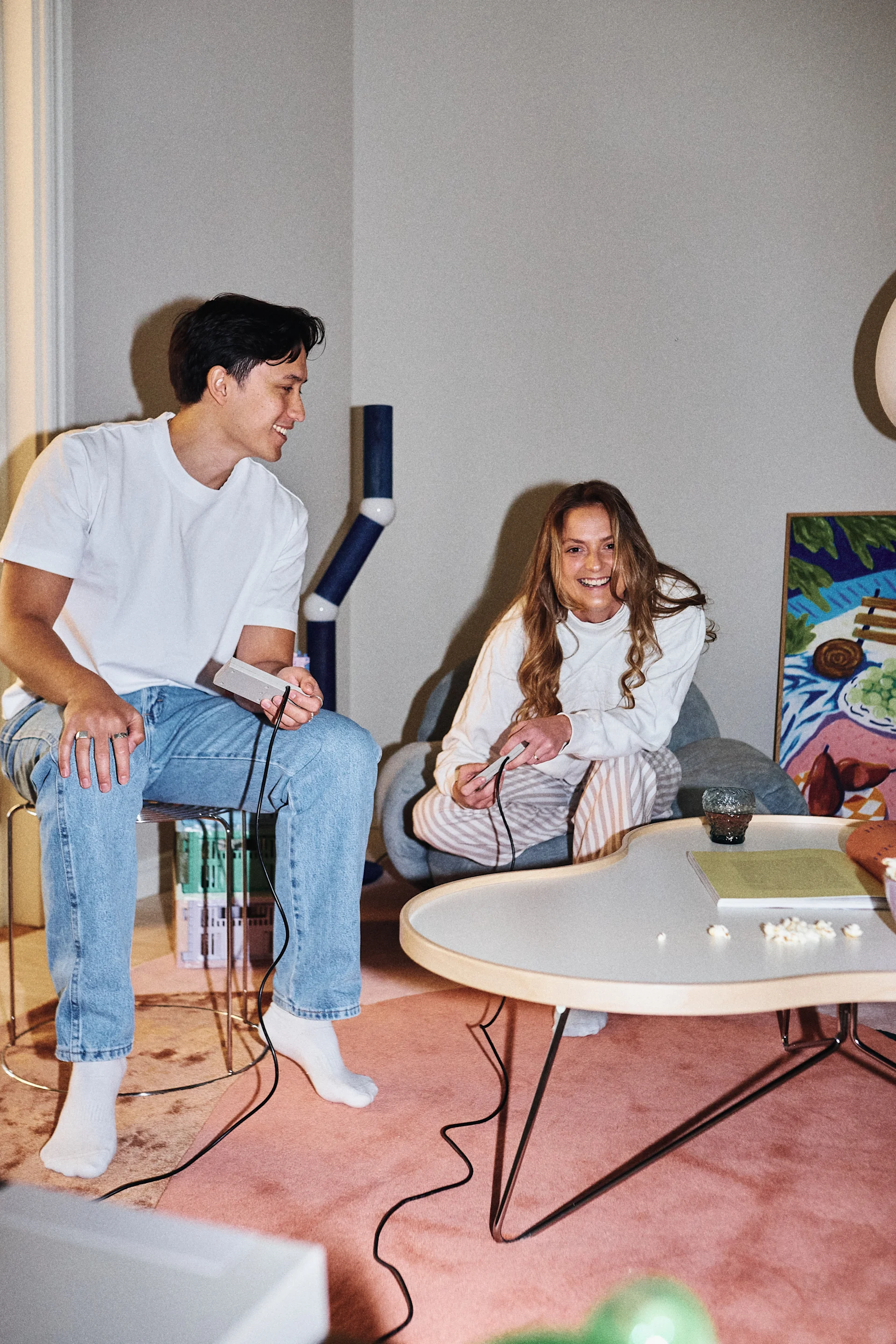 A smiling man and woman playing video games, holding controllers in a comfortable living room with a pink rug.