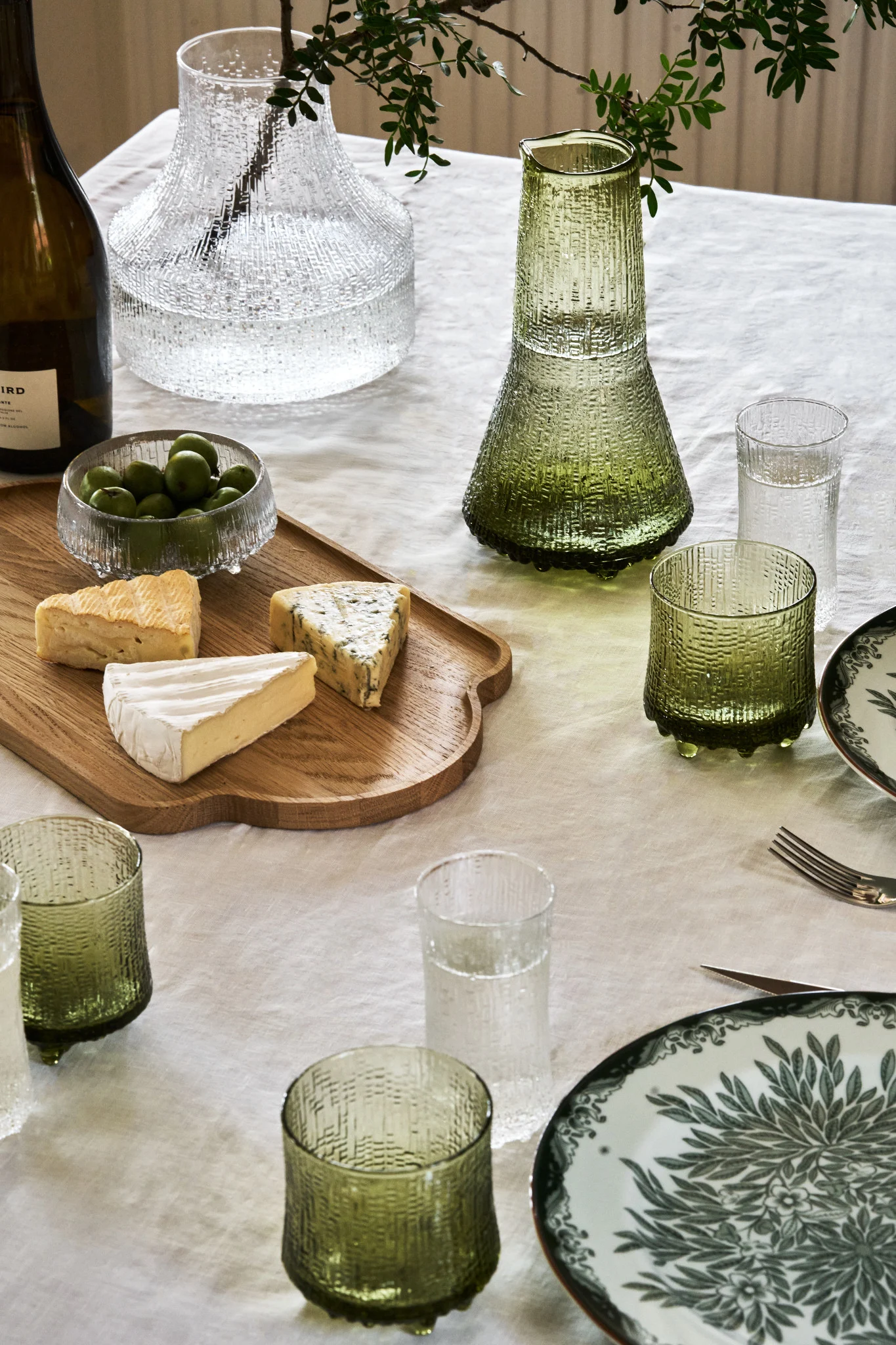 A table set with textured glassware, a wooden board with various cheeses, a bowl of olives, and a patterned plate.