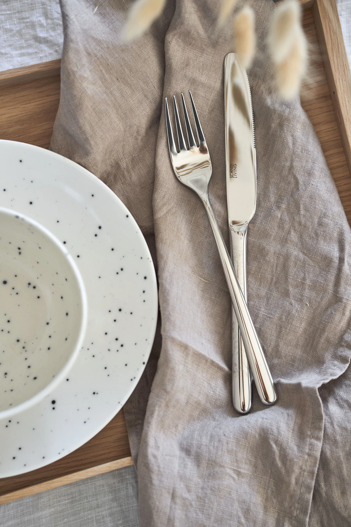 Close-up of a table setting on a wooden tray with a speckled white plate and bowl, a beige linen napkin, and silver fork and knife.