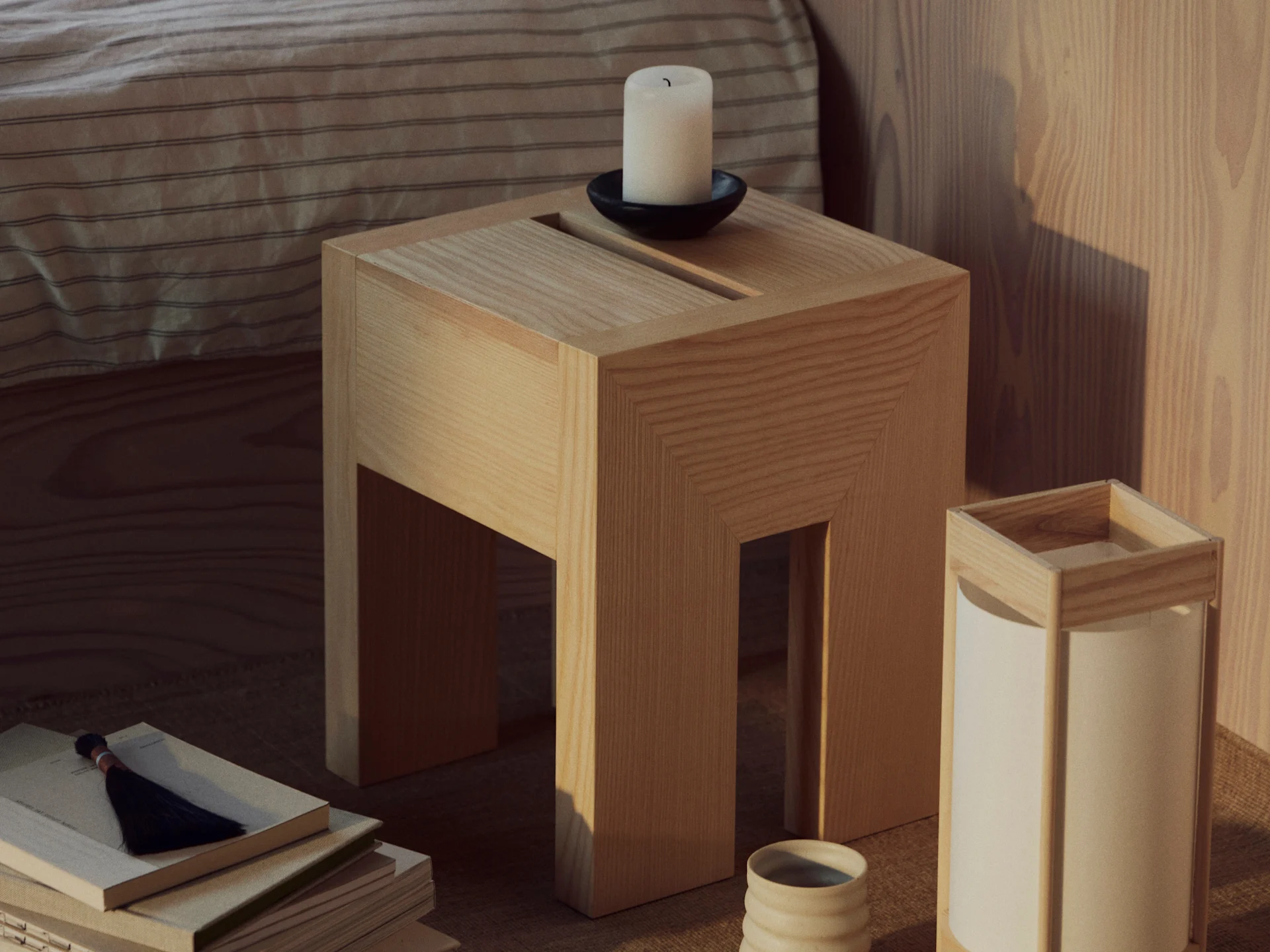 A wooden side table with a white candle in a black holder, a stack of books with a tassel, and a lamp with a ceramic cup.