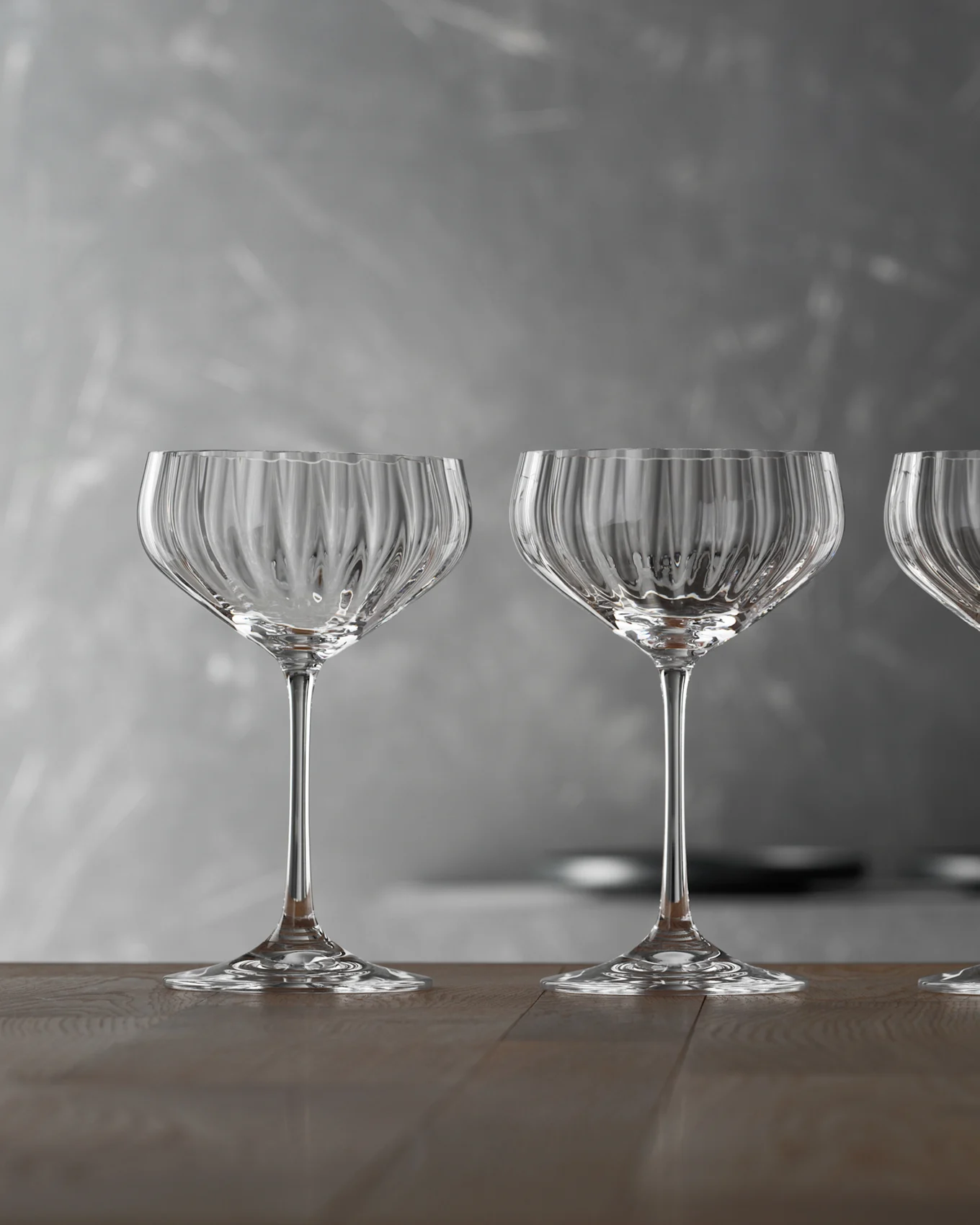 Three empty, clear ribbed glass coupe glasses on a wooden table against a gray background.
