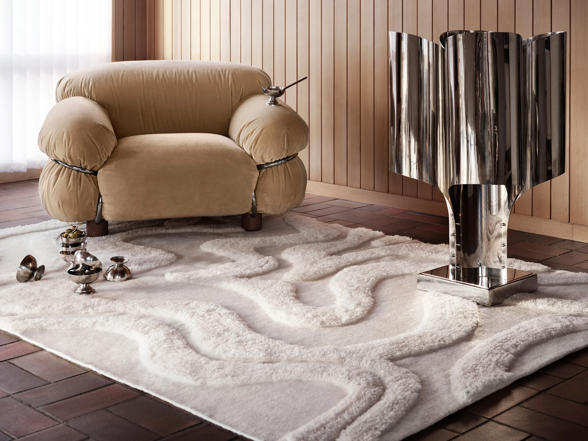 A modern room with a beige, puffy armchair, a tall metallic sculpture, a white rug with wavy patterns, and silver bowls with olives.
