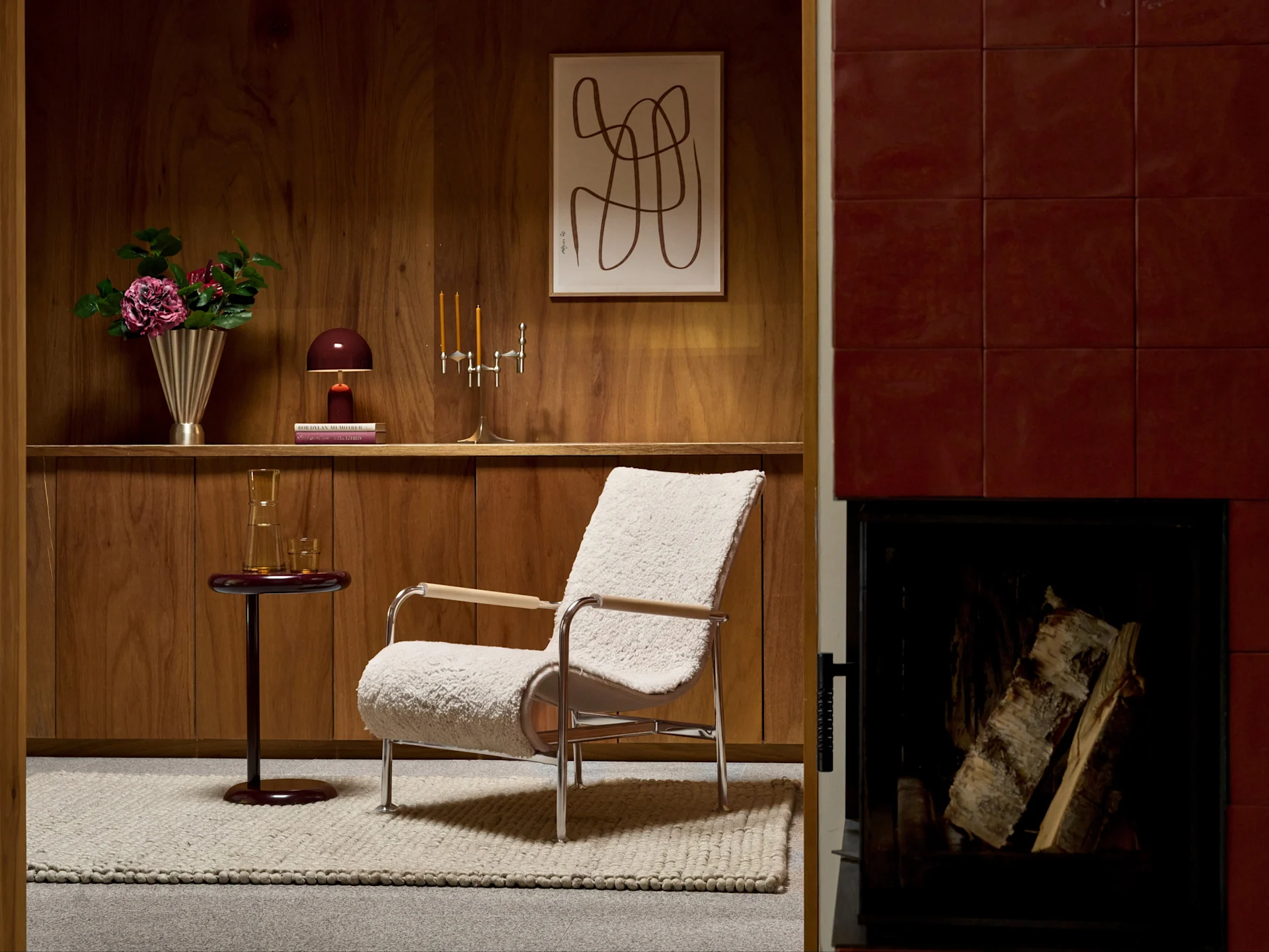A cream bouclé armchair with chrome frame in a wooden paneled room, next to a red tiled fireplace with logs.